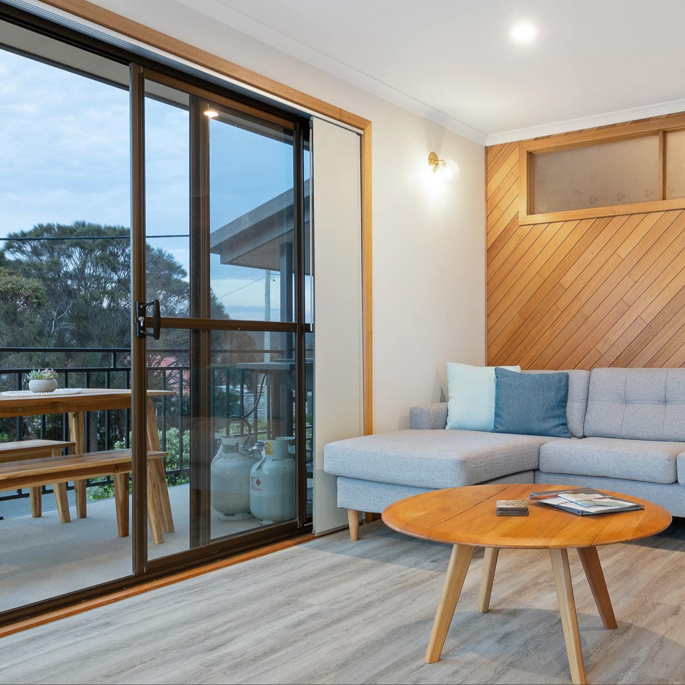 Modern living room with wood-panelled walls, grey sofas, round wooden coffee table, glass door to attached balcony.