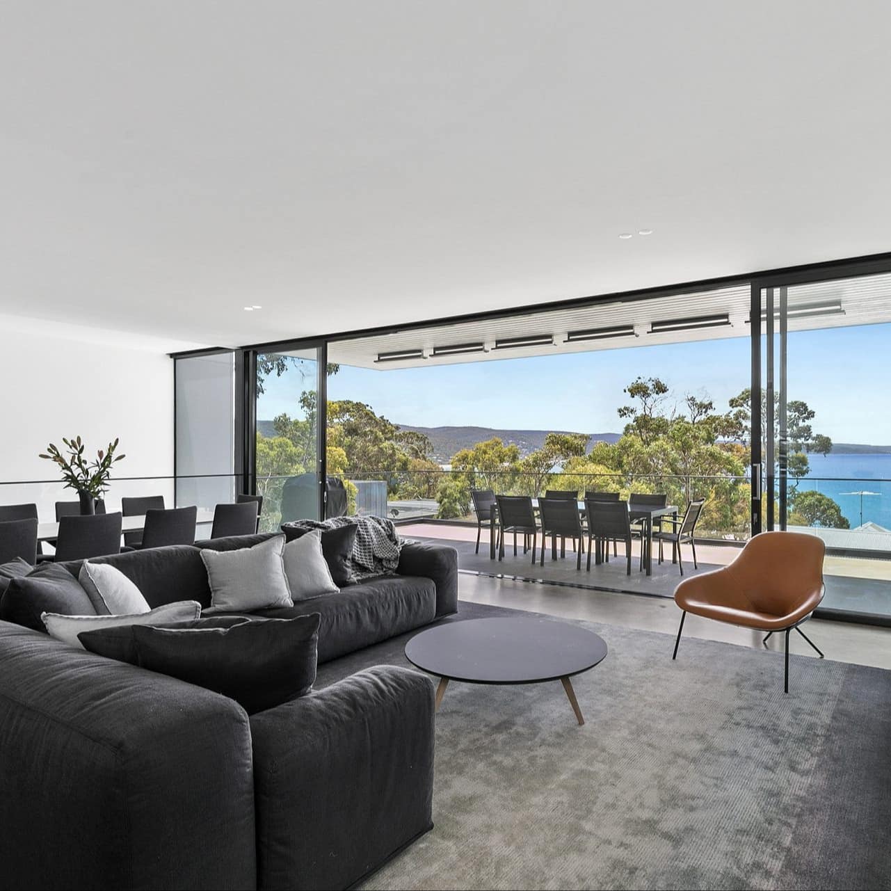 Open living and dining room, cement floors, black sectional, floor-to-ceiling glass doors with panoramic ocean views.