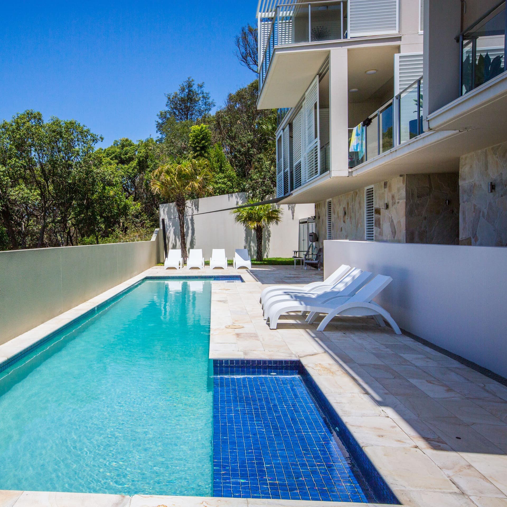 A long pool with a beige pool deck and white lounge chairs, enclosed by beige stone wall, overlooking lush greenery.