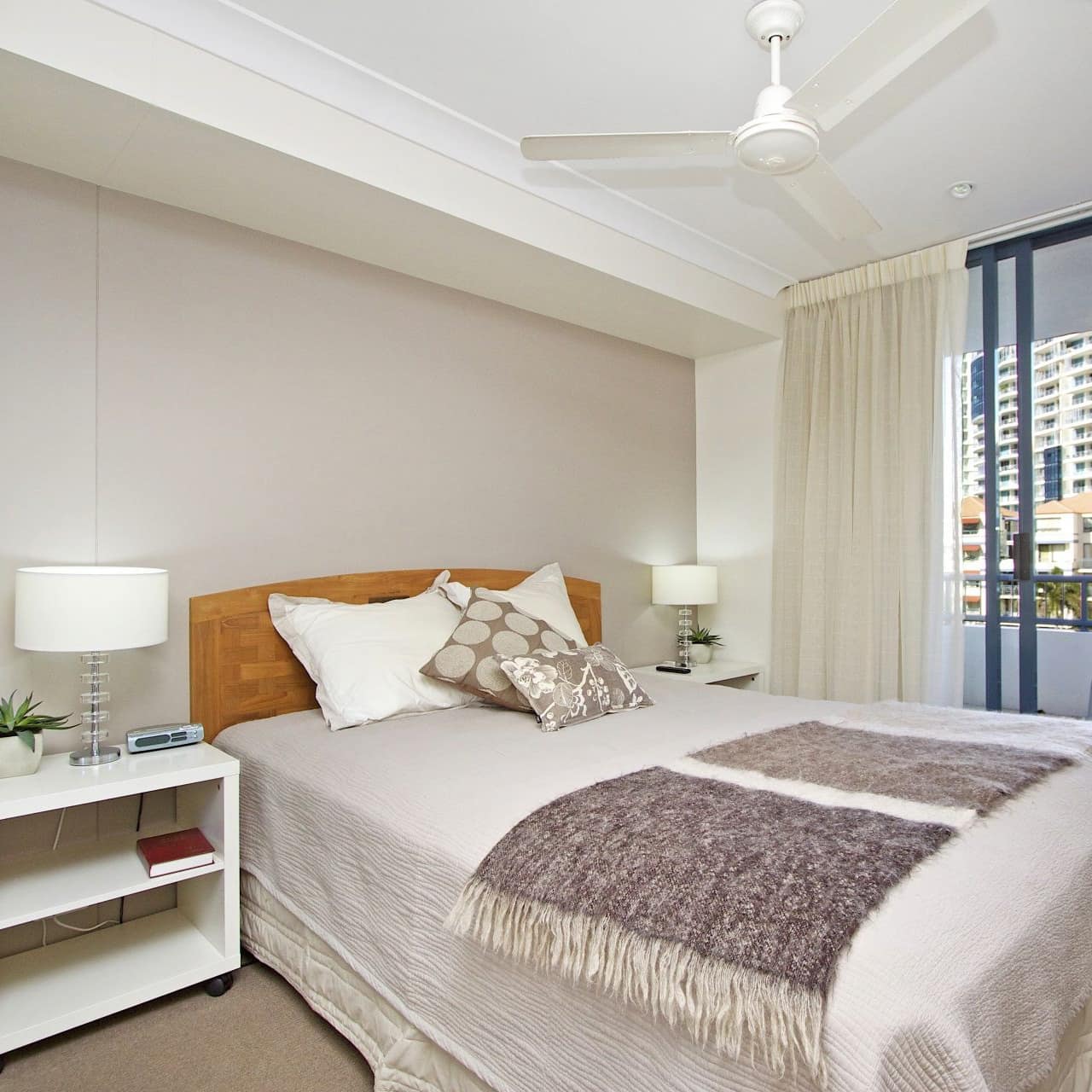 Bedroom with attached balcony, queen size bed, beige bedding, matching white bedside tables, beige walls and carpet.