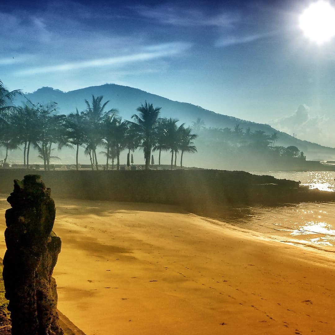 A bright afternoon sun shines down upon a golden beach at low tide with palms and a mountain in the background