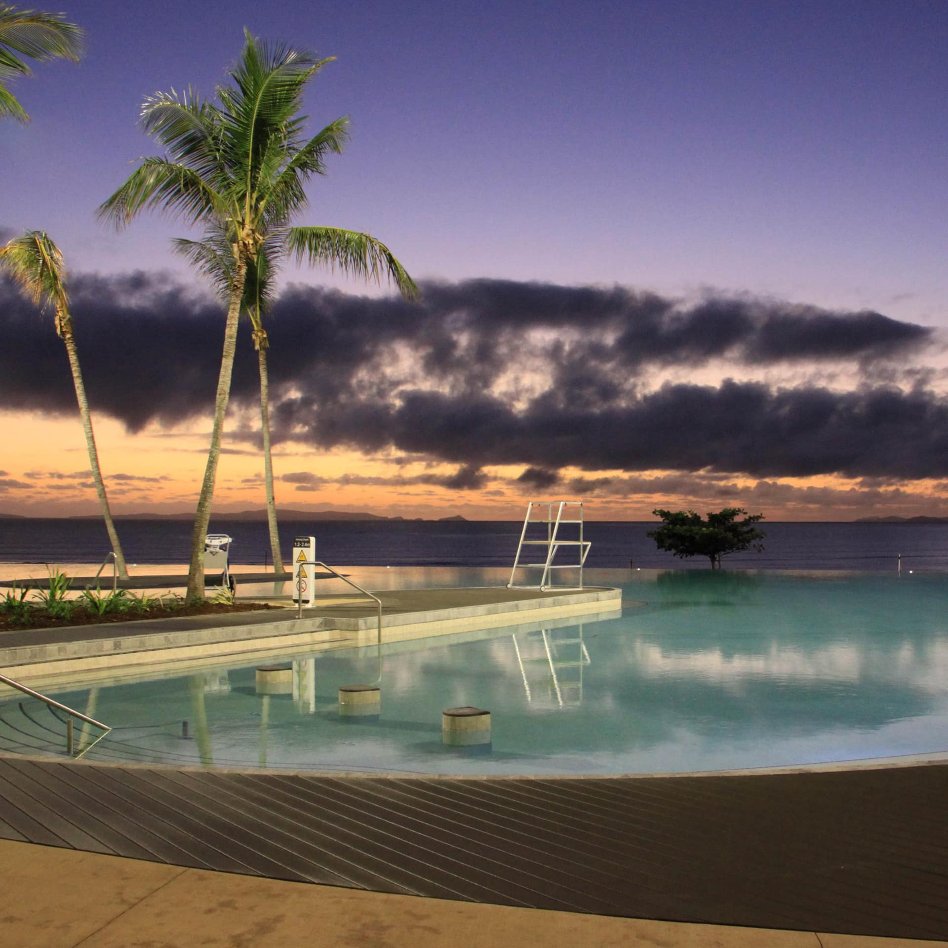 Serene view of a pool at sunset, palm trees around the pool and the ocean visible along the horizon.