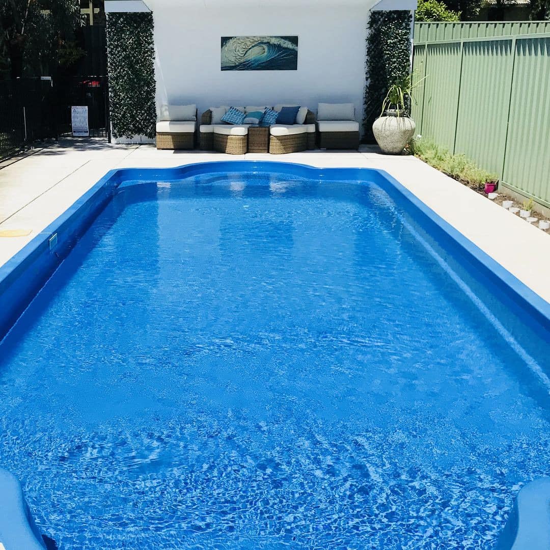 View of crystal blue, inground pool with modern wicker outdoor seating, white cement patio space, and potted plants.