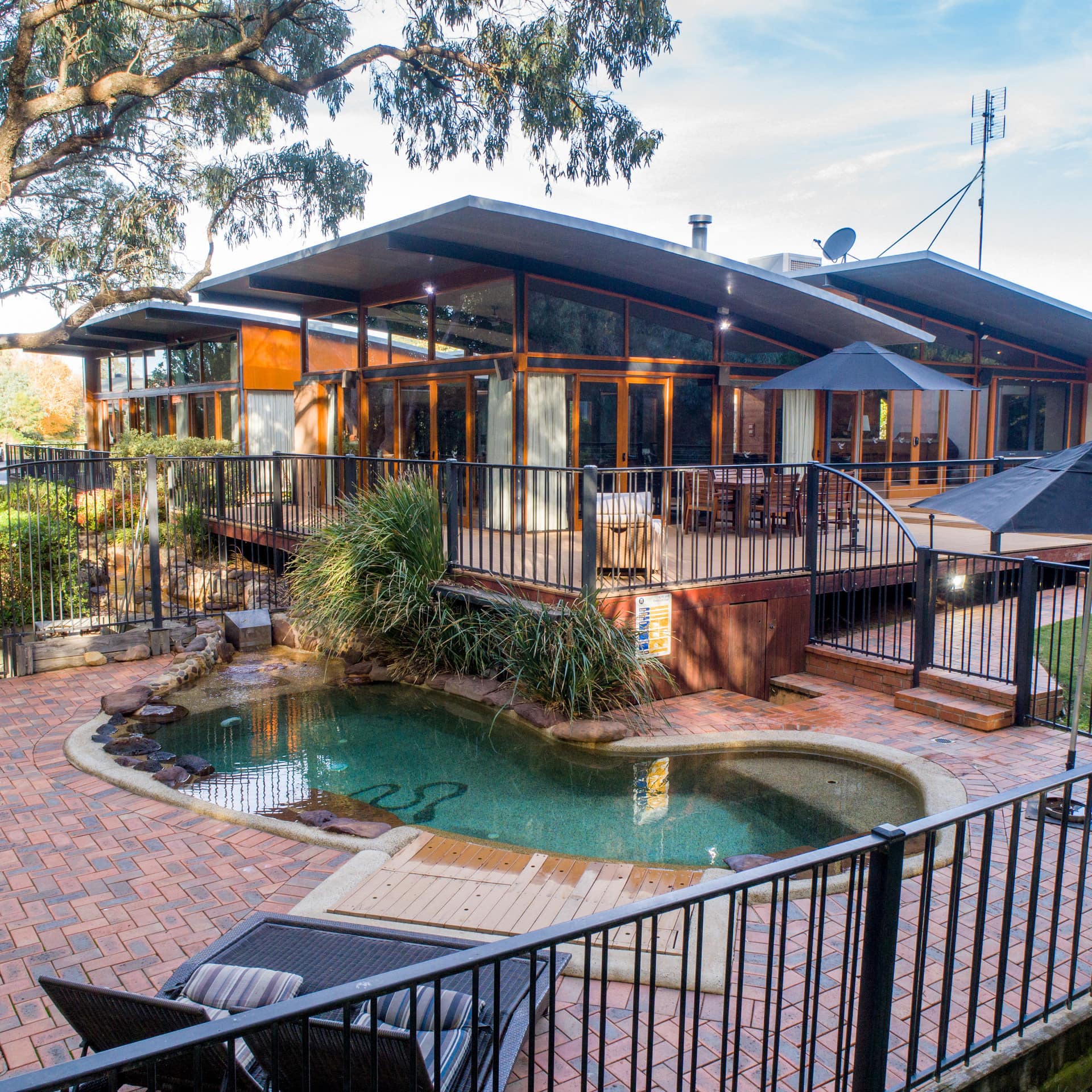 Exterior view of modern home with a private pool, brick pool deck, wrap-around deck with dining set, and a manicured lawn.