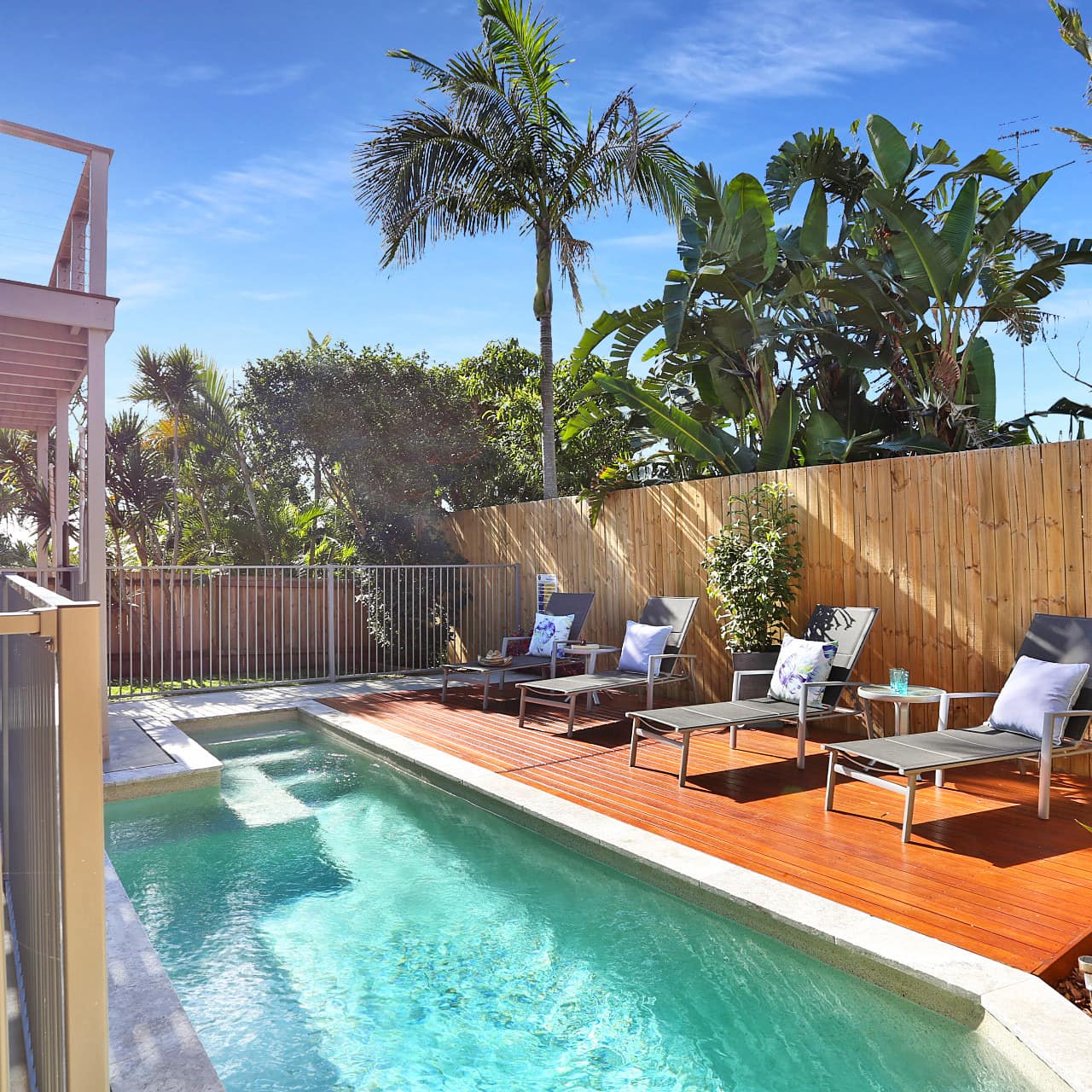 Sunny view of a pool with wooden pool deck and black lounge chairs enclosed by a wooden fence and palm trees.