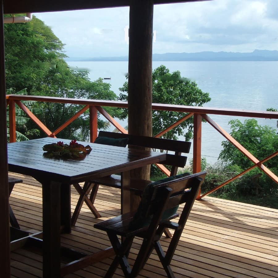 A wooden deck with a wooden dining set, overlooking the ocean lush greenery.