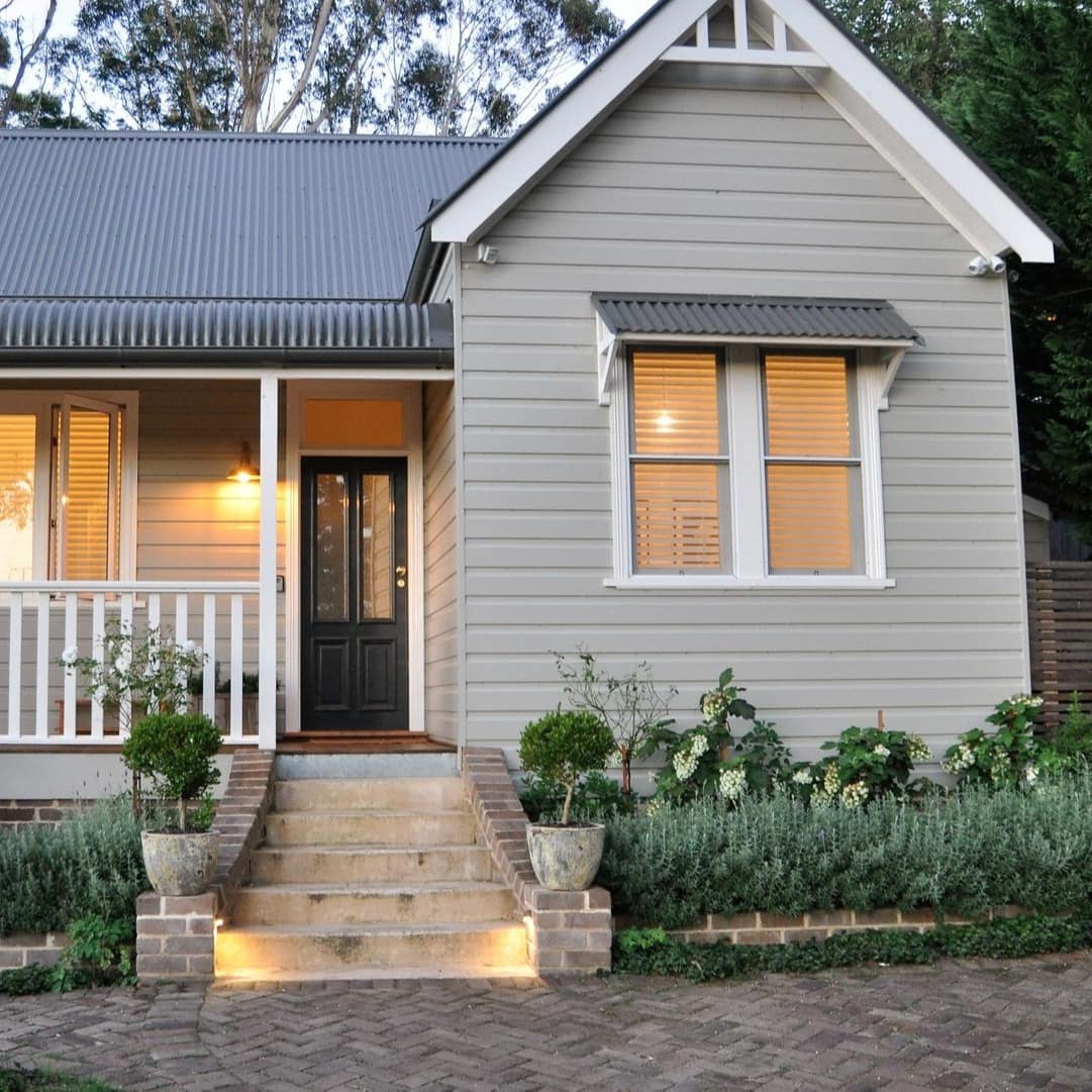 Exterior of cottage-style home with beige panel siding, white trim and metal roof, brick planters and stairs to front porch.