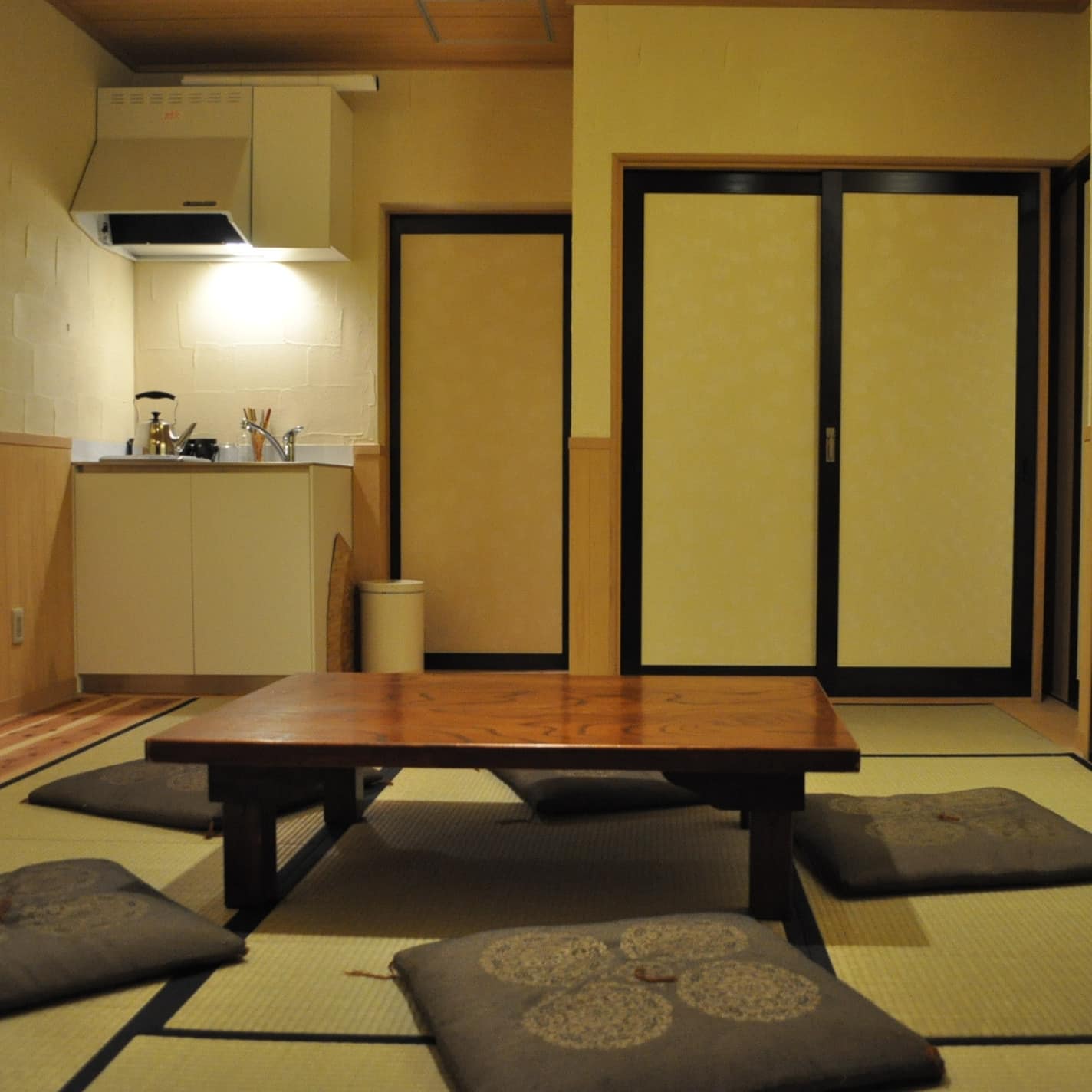 A room with tatami floors, sliding doors, a short wooden table with floor cushions, and a kitchenette.