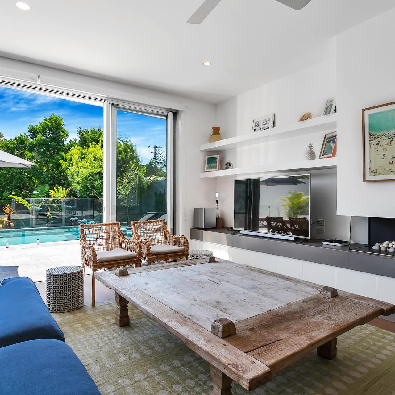 A modern living room with blue sofa, rattan chairs, reclaimed wood table, and private pool through the sliding glass doors.