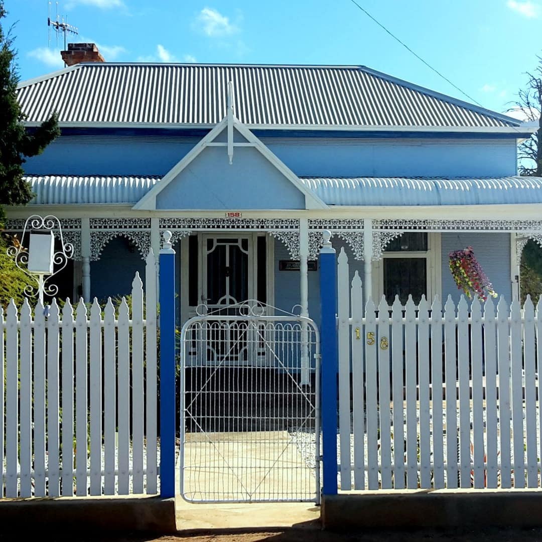 Exterior of a heritage miner’s cottage with bright blue paint, intricate trim on the porch, and a white picket fence.