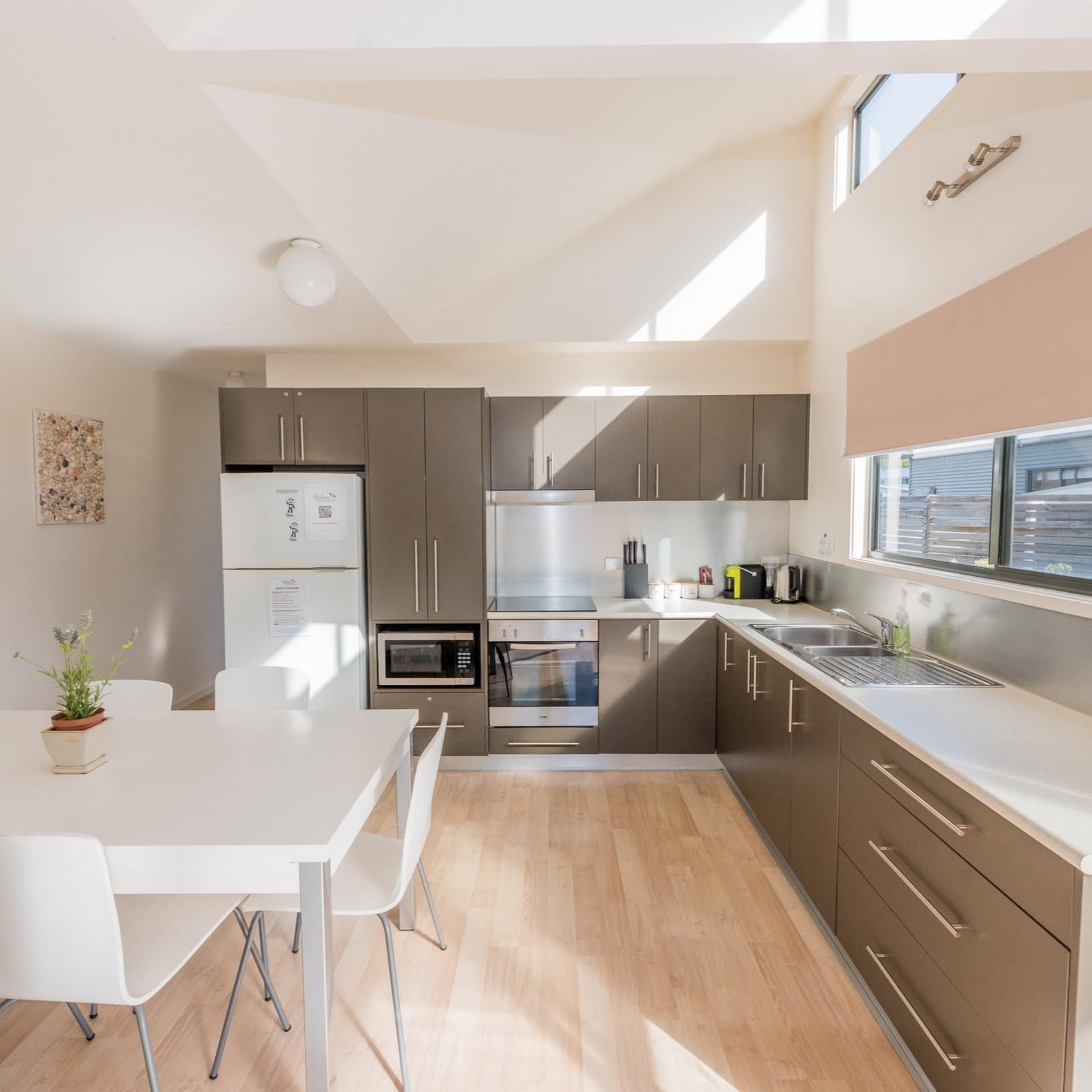 Sunny, modern kitchen with white dining set, grey cabinets, white countertops, and wide windows.