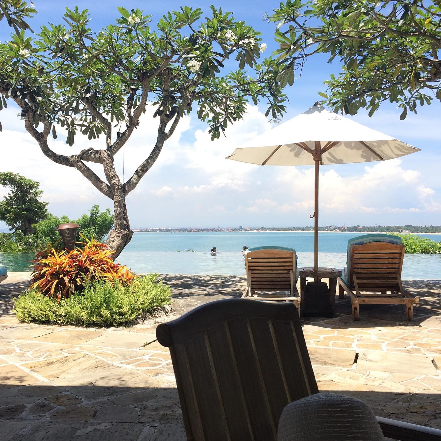 Stone patio of waterfront villa with wood lounges, a shade umbrella and trees against an infinity pool