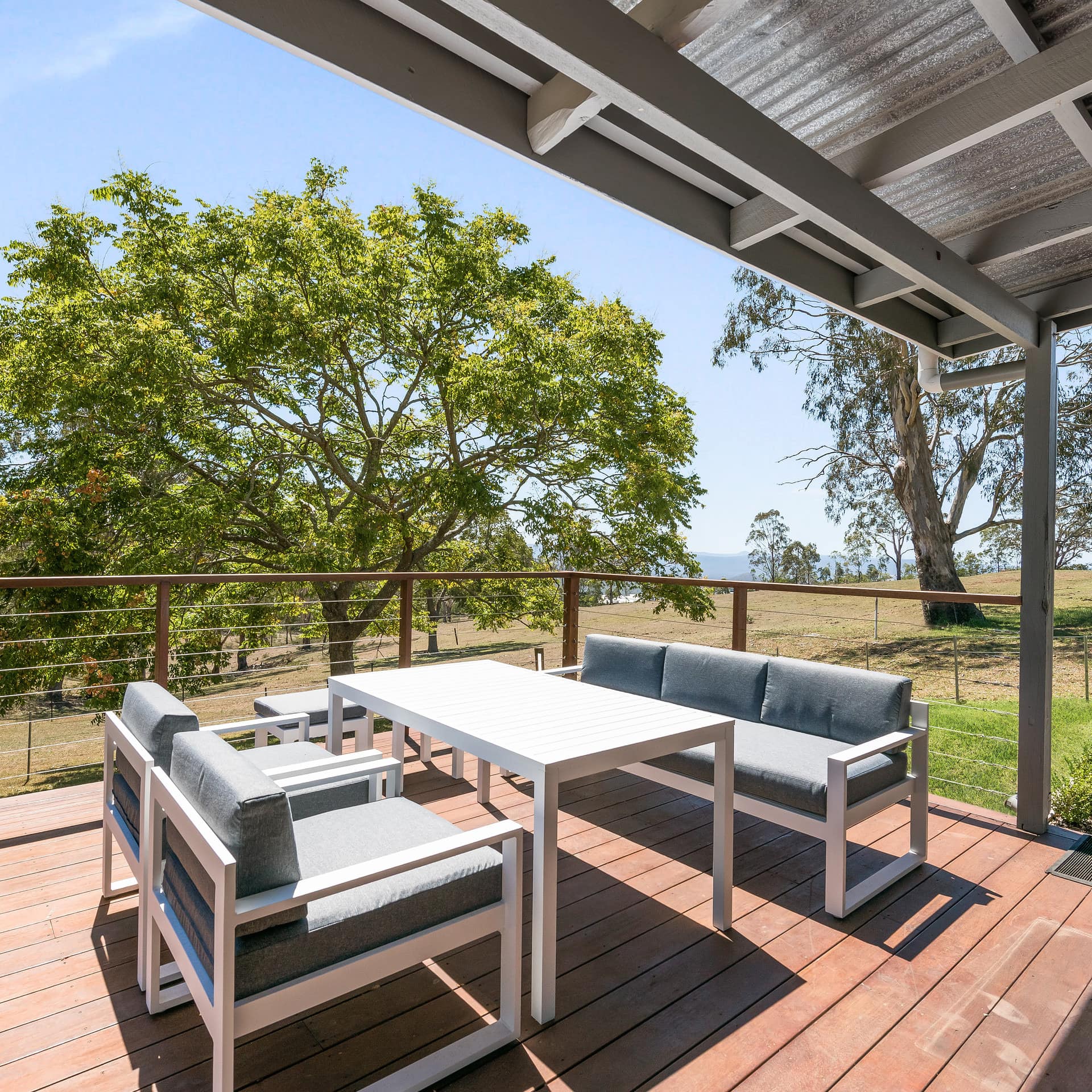 A partially covered deck with a white dining table and grey cushioned chairs and sofa, surrounded by rural landscape.