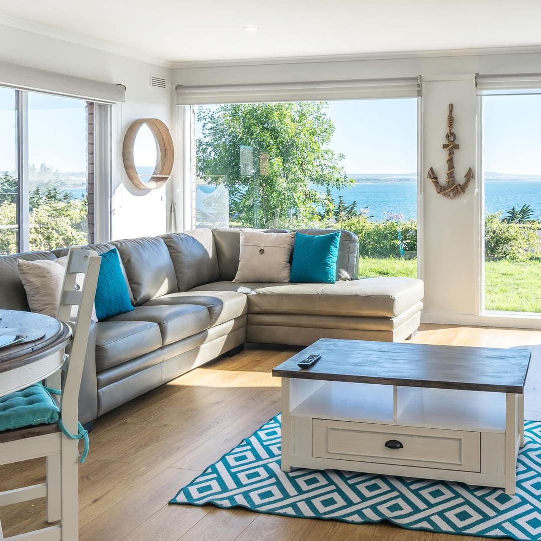 Open living and dining room with teal and white decor, grey leather sectional, floor to ceiling windows with ocean views.