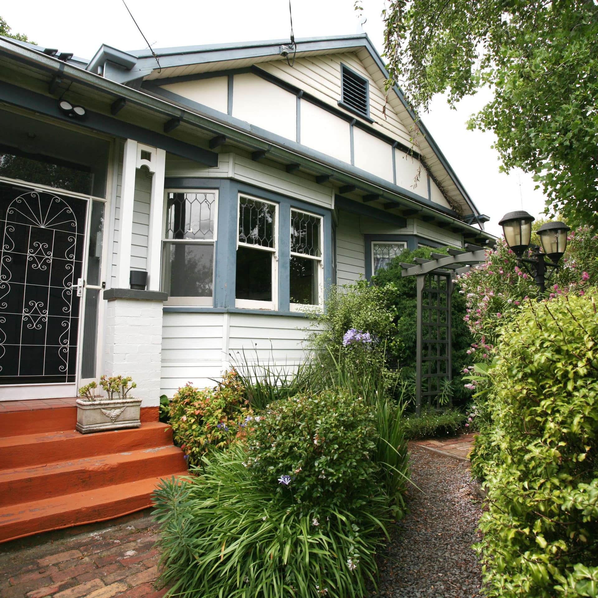 Charming white cottage with blue trim, orange stairs to a front door, pebble gravel path, and a lush garden.