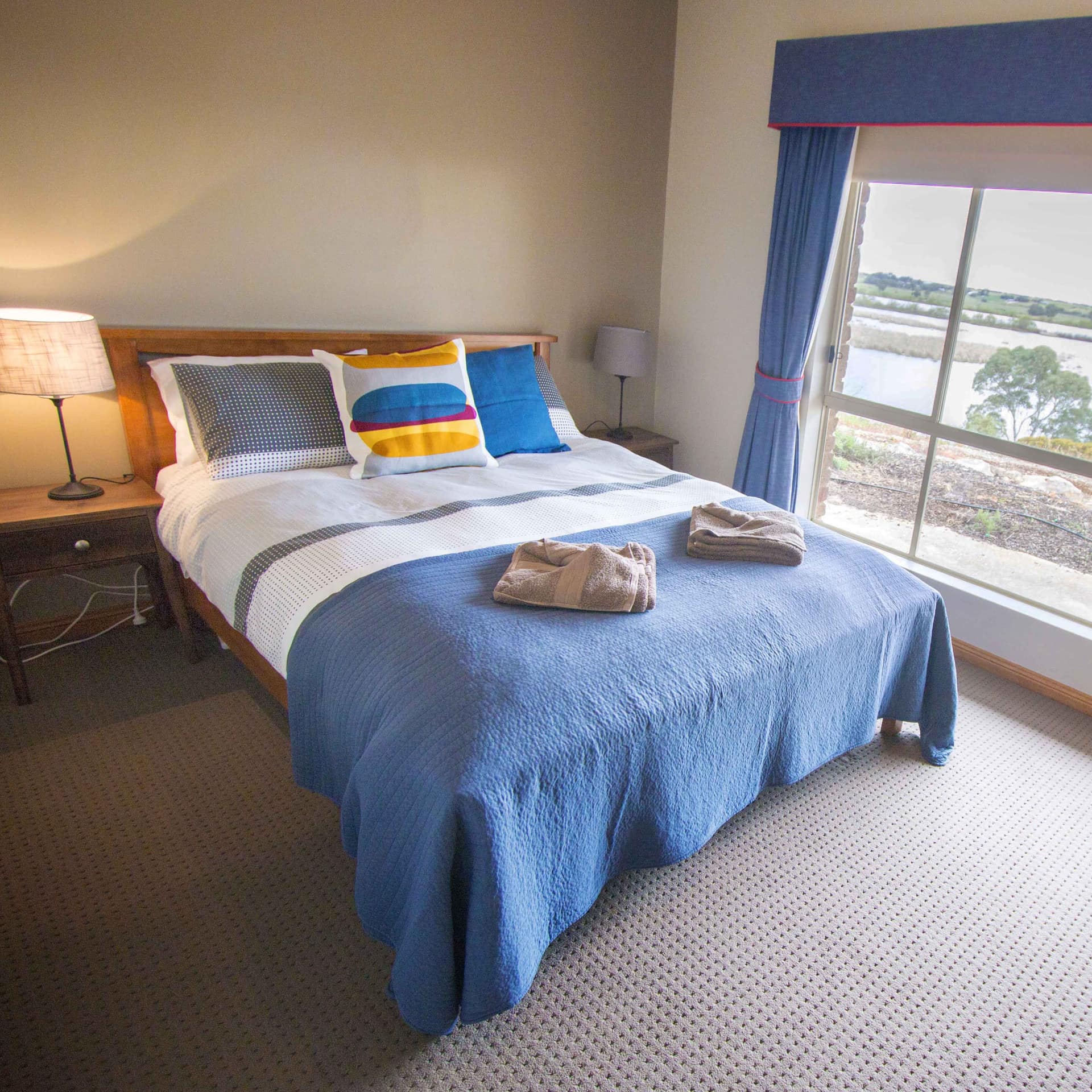 Bedroom with grey carpet, a wood-framed king-size bed, wide windows with lake views and blue curtains.