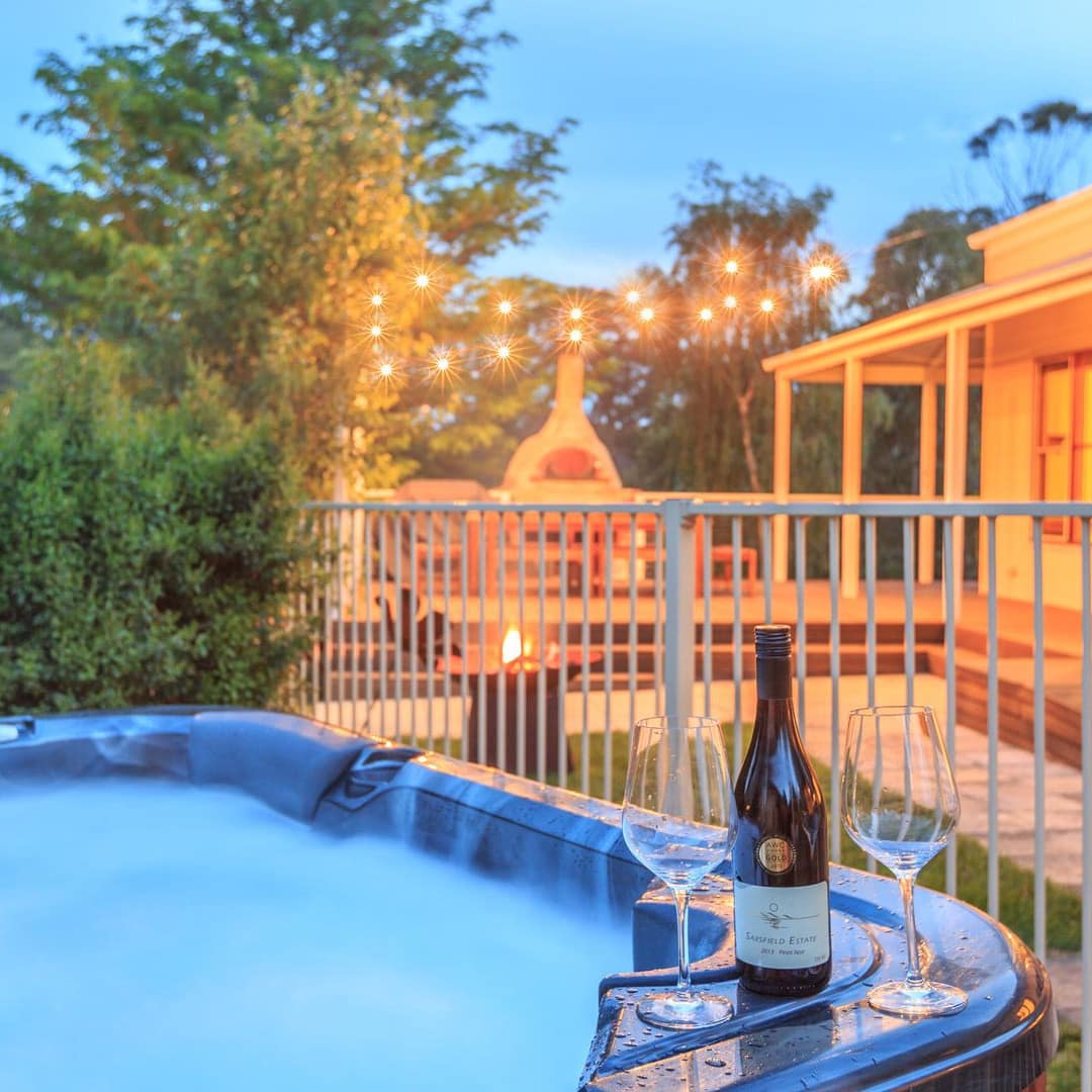Wine glasses sitting on the edge of a hot tub in a charming backyard with outdoor kitchen and hanging lights.