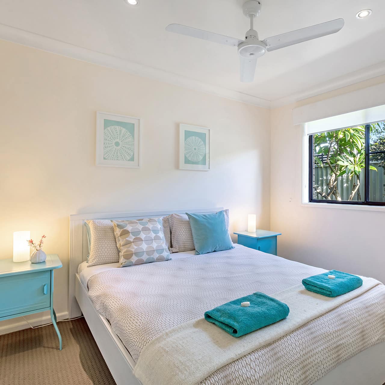 Bedroom with teal and white decor, queen size bed, beige carpet, white walls, and a window with views of tropical plants.