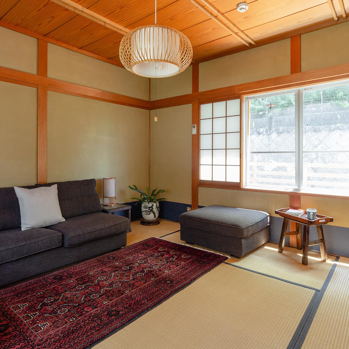 Traditional Japanese room with tatami floors, grey sectional, red area rug, woven bamboo hanging light and wide windows.