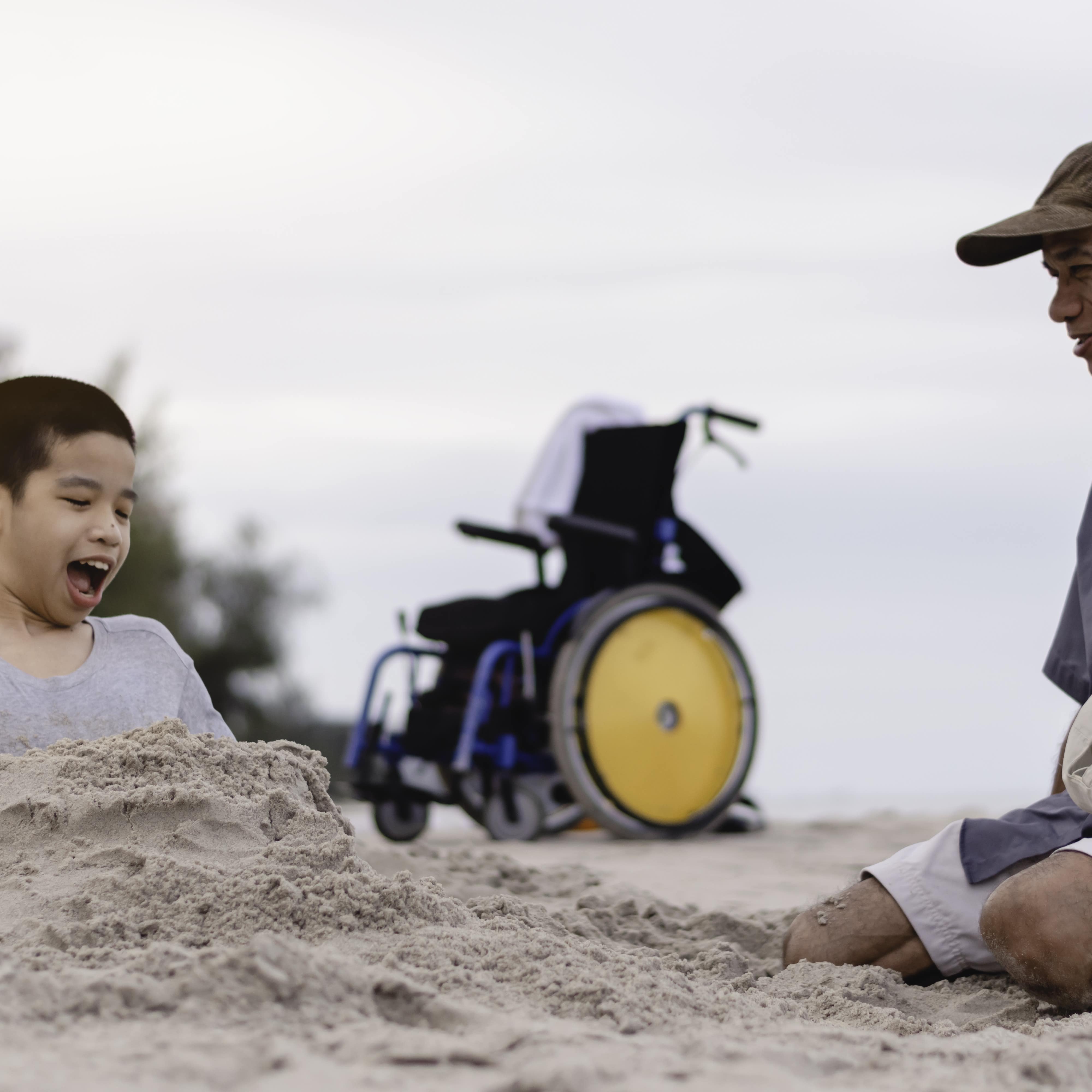 Young boy covered in sand with wheelchair in background and his father on the beach