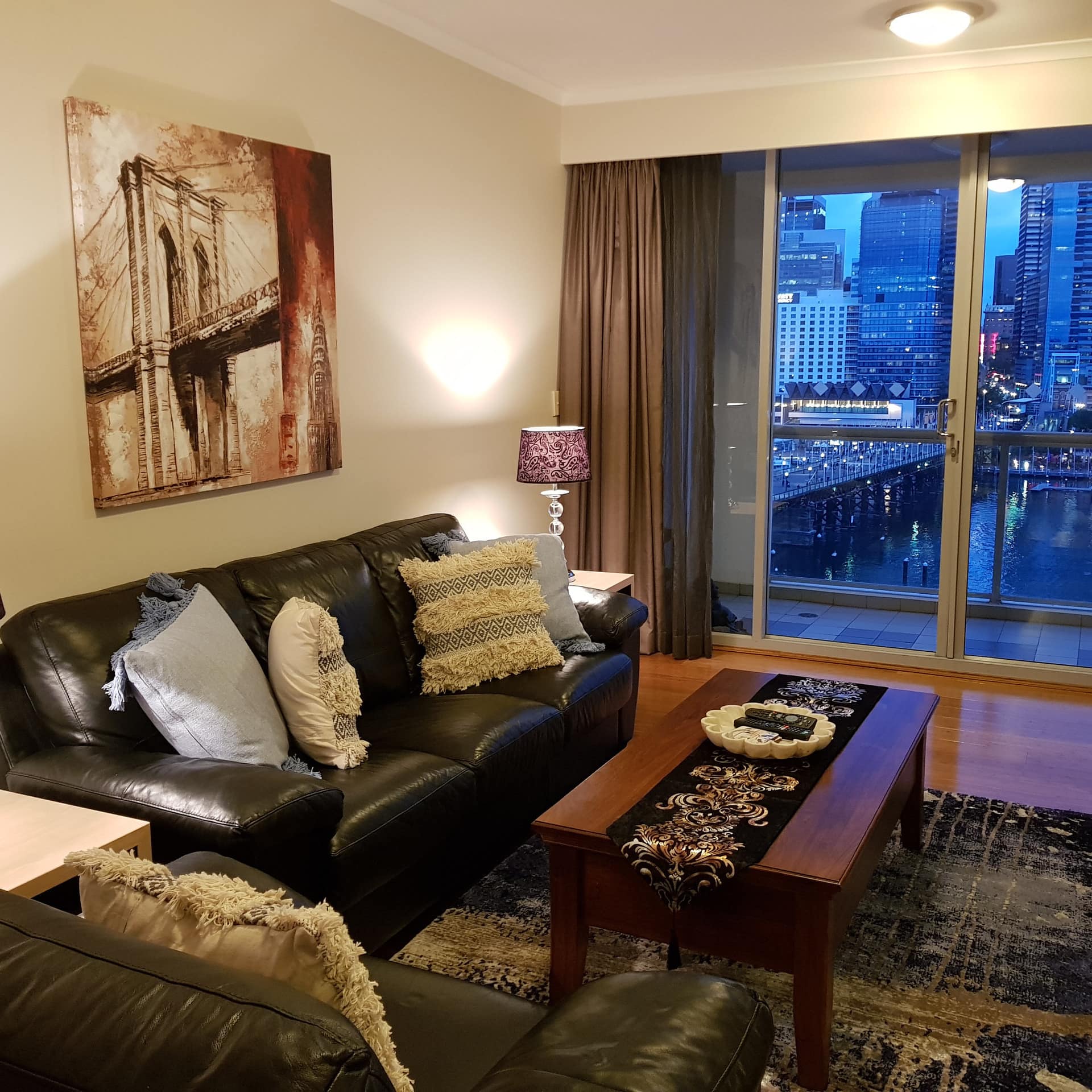 An open living room with black and brown furnishings, and a balcony with a nighttime view of the city and Darling Harbour.