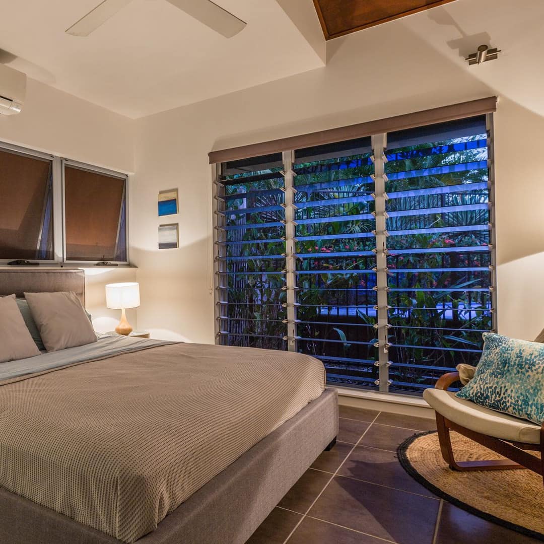 A bedroom with tan tile floors and beige walls, a king size bed with tan bedding, white canvas chair, tall vented window.