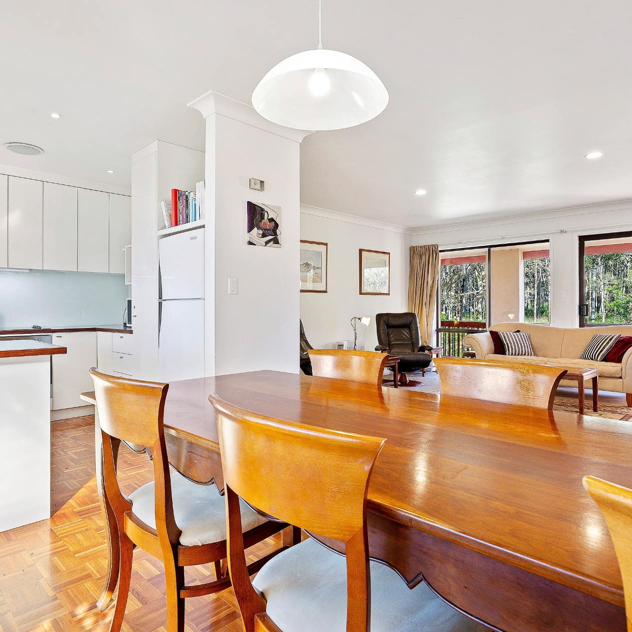 Open floor plan living, dining and kitchen area with a long teak table, white kitchen cabinetry, and classic roll-arm sofa.