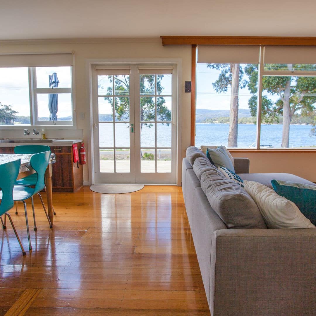Open living and dining room, wooden dining table with teal chairs, grey sofa, wide windows with ocean views.