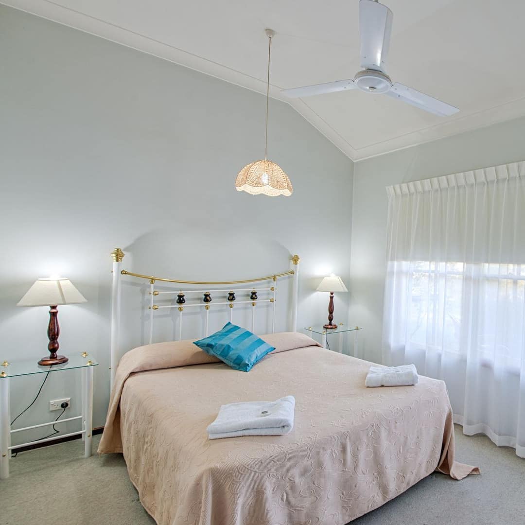 Spacious bedroom, white walls and beige carpet, queen size bed, light pink bedding, wide windows with sheer curtains.