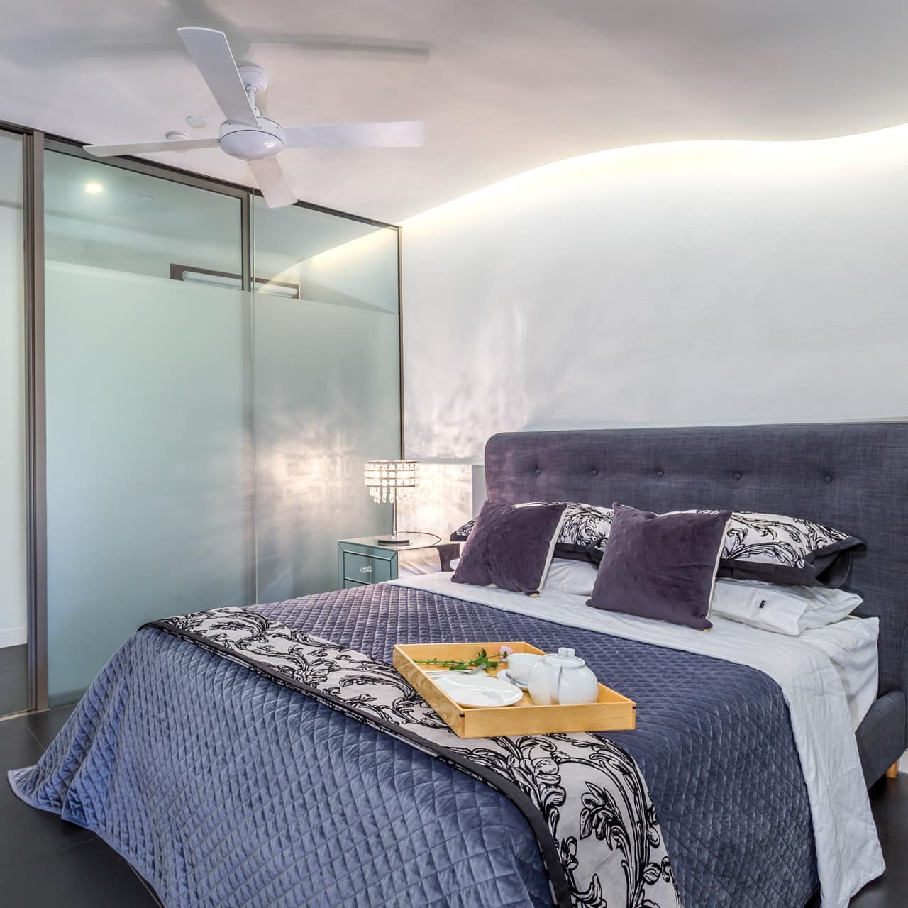 A bedroom with grey tile floors, white walls, a king size bed, metallic purple bedding, and a tray set with tea.