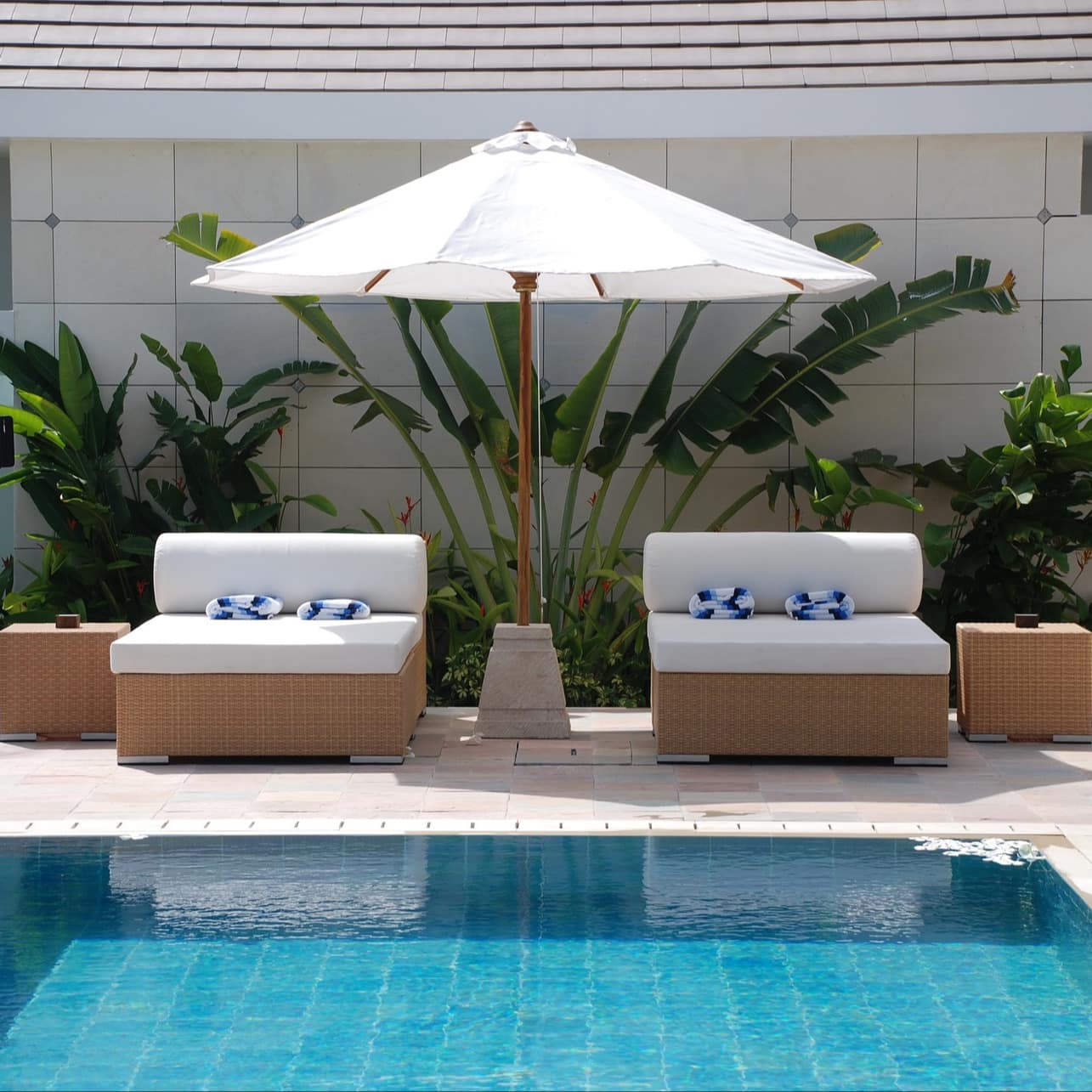 Private pool of Seminyak villa with turquoise water, two low-back wicker chairs, a shade umbrella and tropical plants