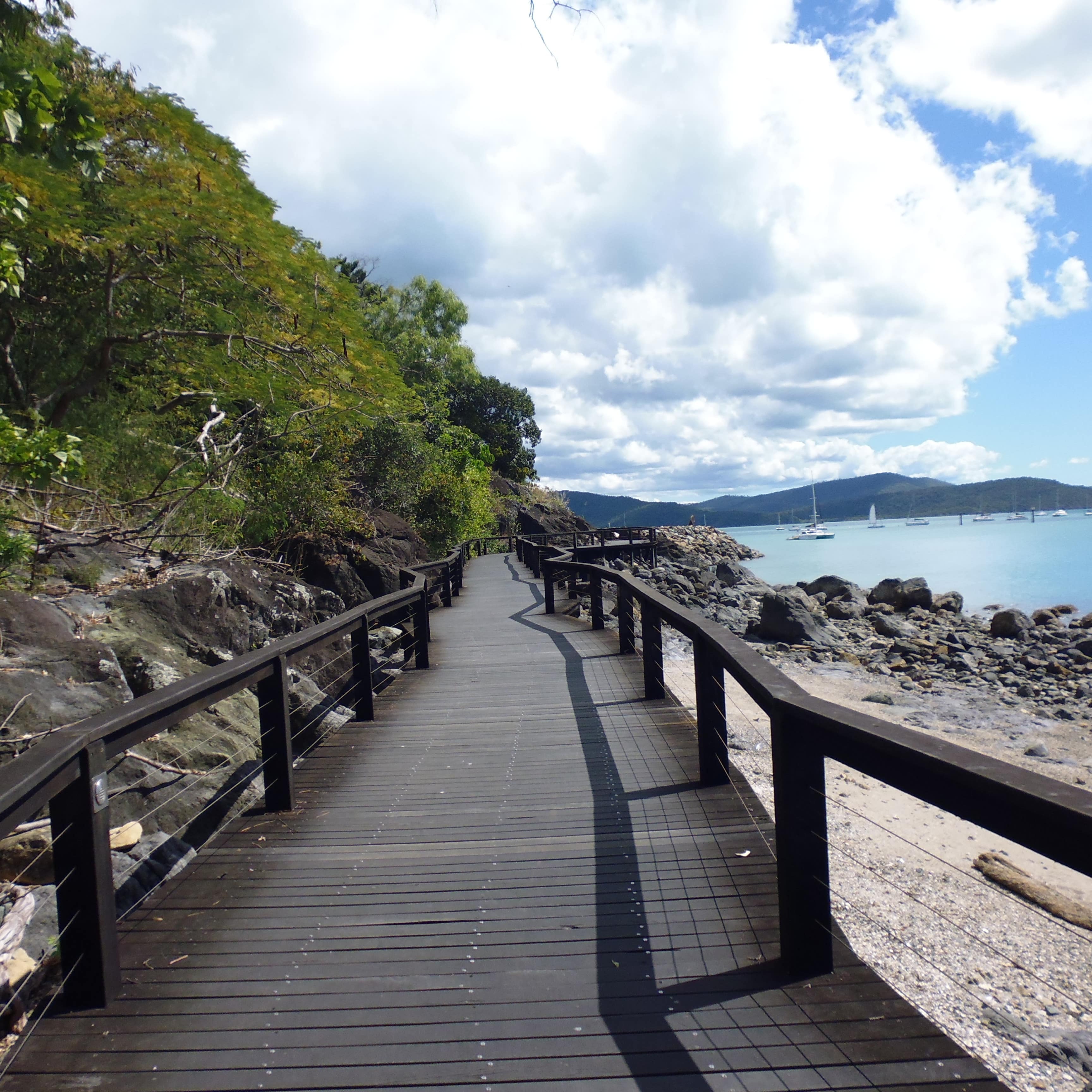 Wooden oceanside walkway with railings, trees on the hillside and boulders down to the shore with sailboats in the background