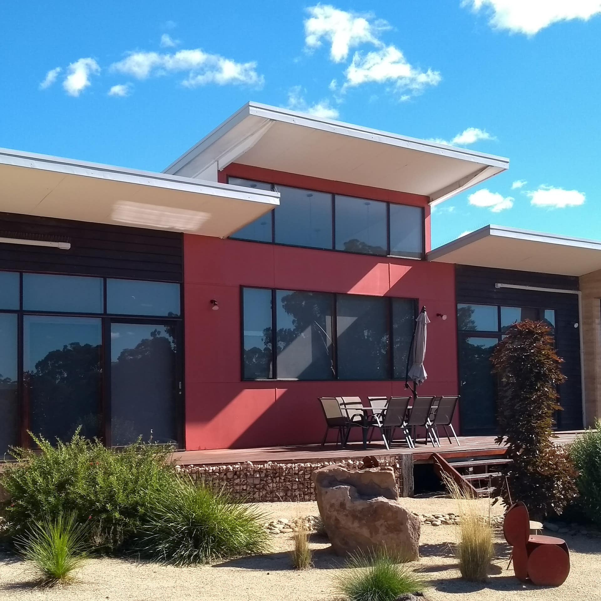Large modern home with tall windows and red siding, a covered porch with a dining set, and a landscaped yard.