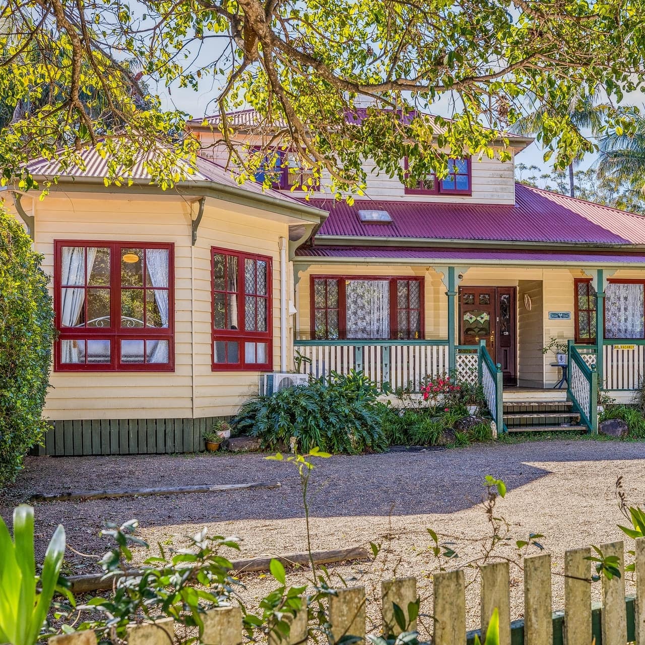 Exterior view of yellow cottage with red trim and roof, green railing on the covered front porch, lush vegetation in yard.