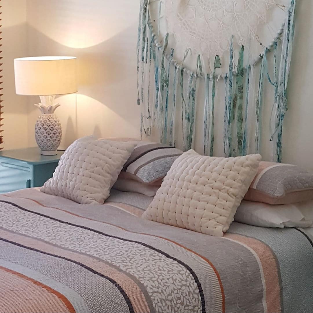 Bedroom with white walls, queen size bed, colorful bedding, woven wall hanging, teal bedside tables, tall windows.