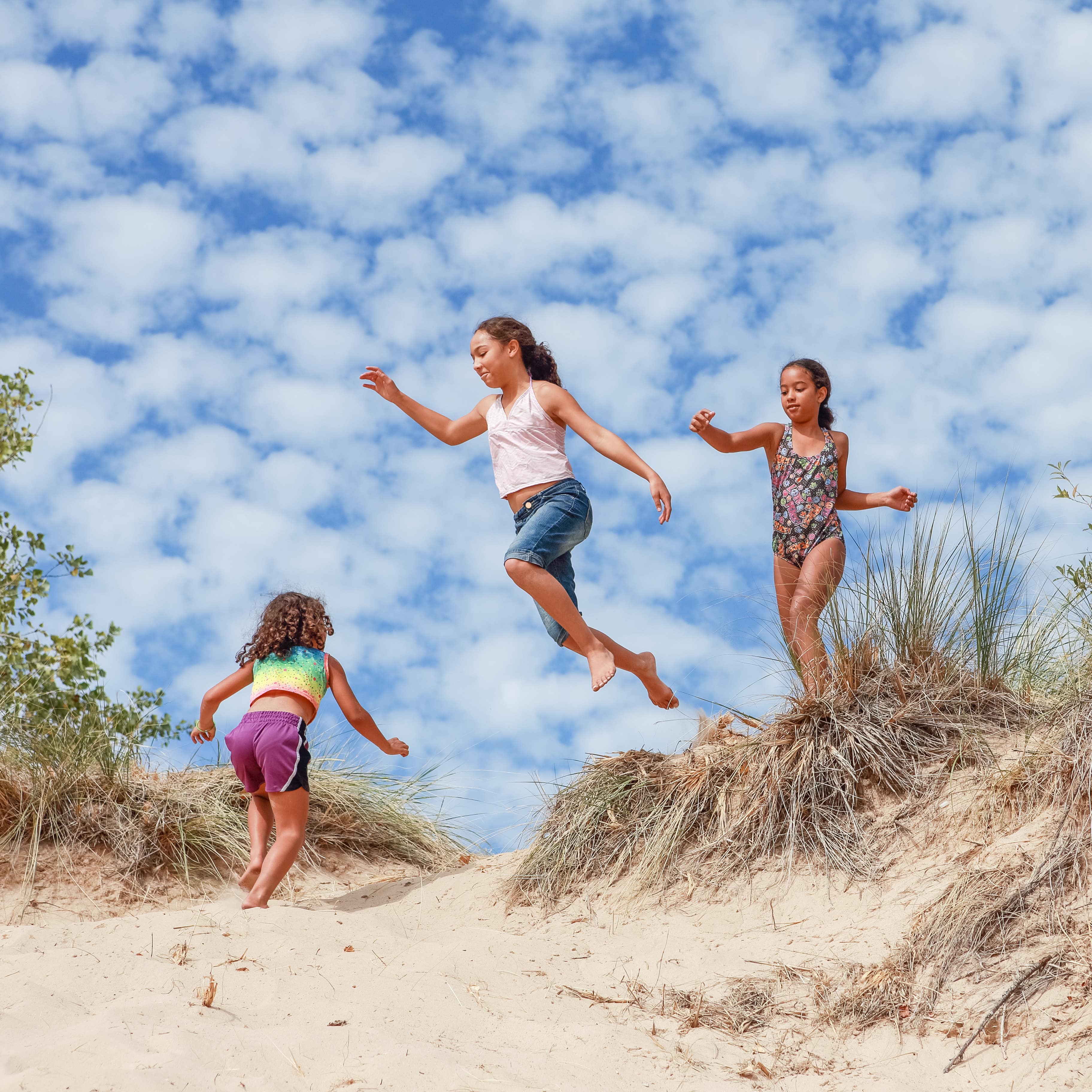Three children jumping from sand dunes at beach against blue cloudy sky