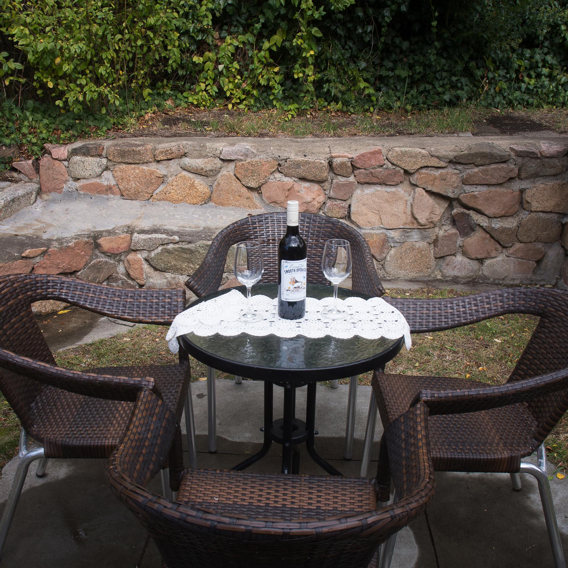 Sunken terrace area with natural stone walls, brown wicker dining set, lace table runner and wine glasses set up on table.