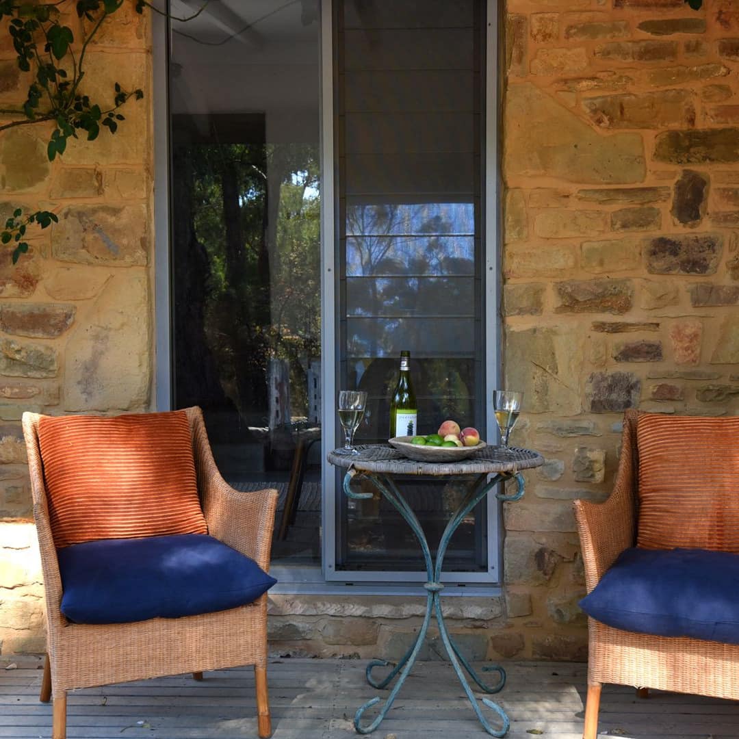 View of two wicker chairs and metal table with wine and fruit on a wooden deck, the home has natural stone siding.