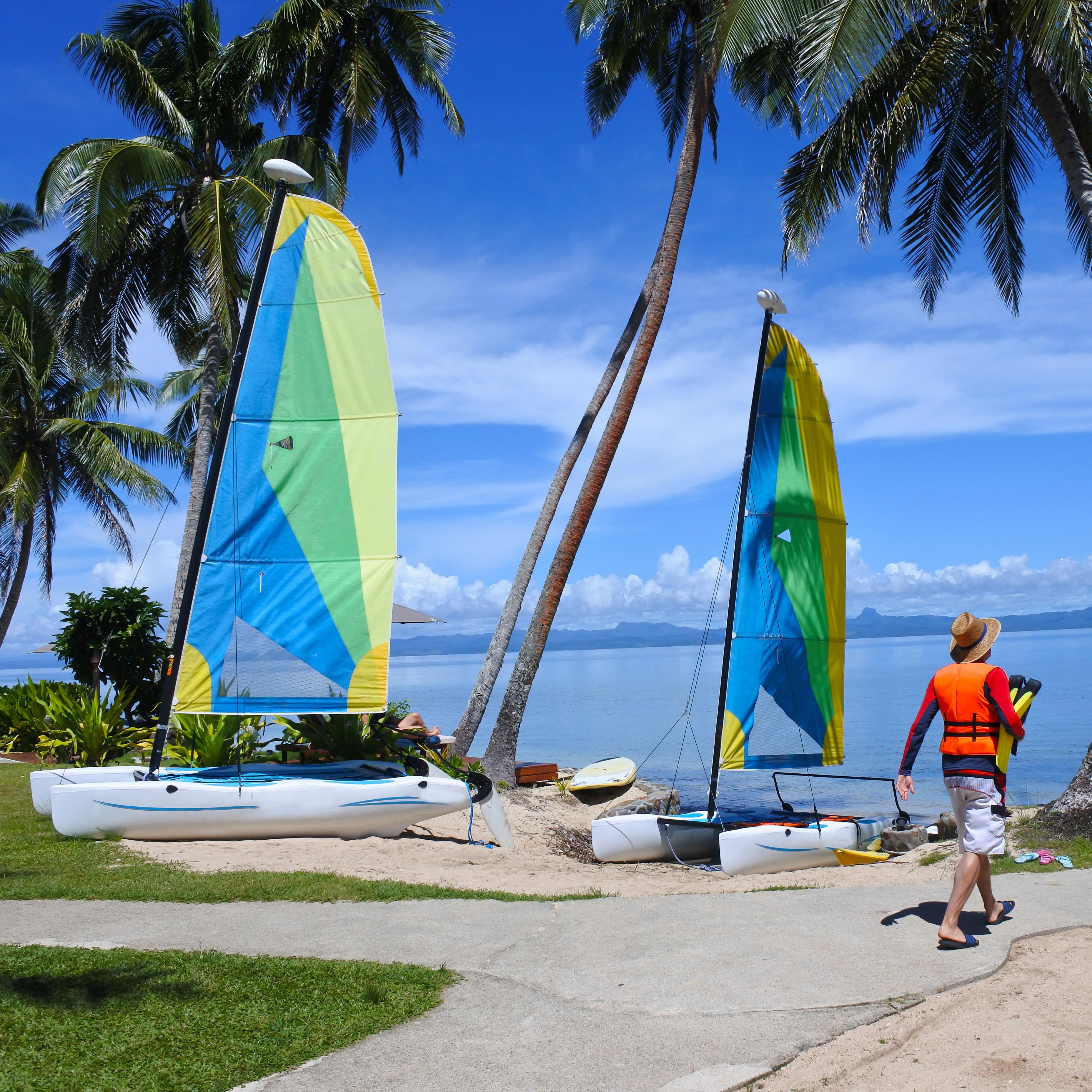 Tourist walks toward sea getting ready to sail at Fiji resort beach with catamarans and palm trees in the sand