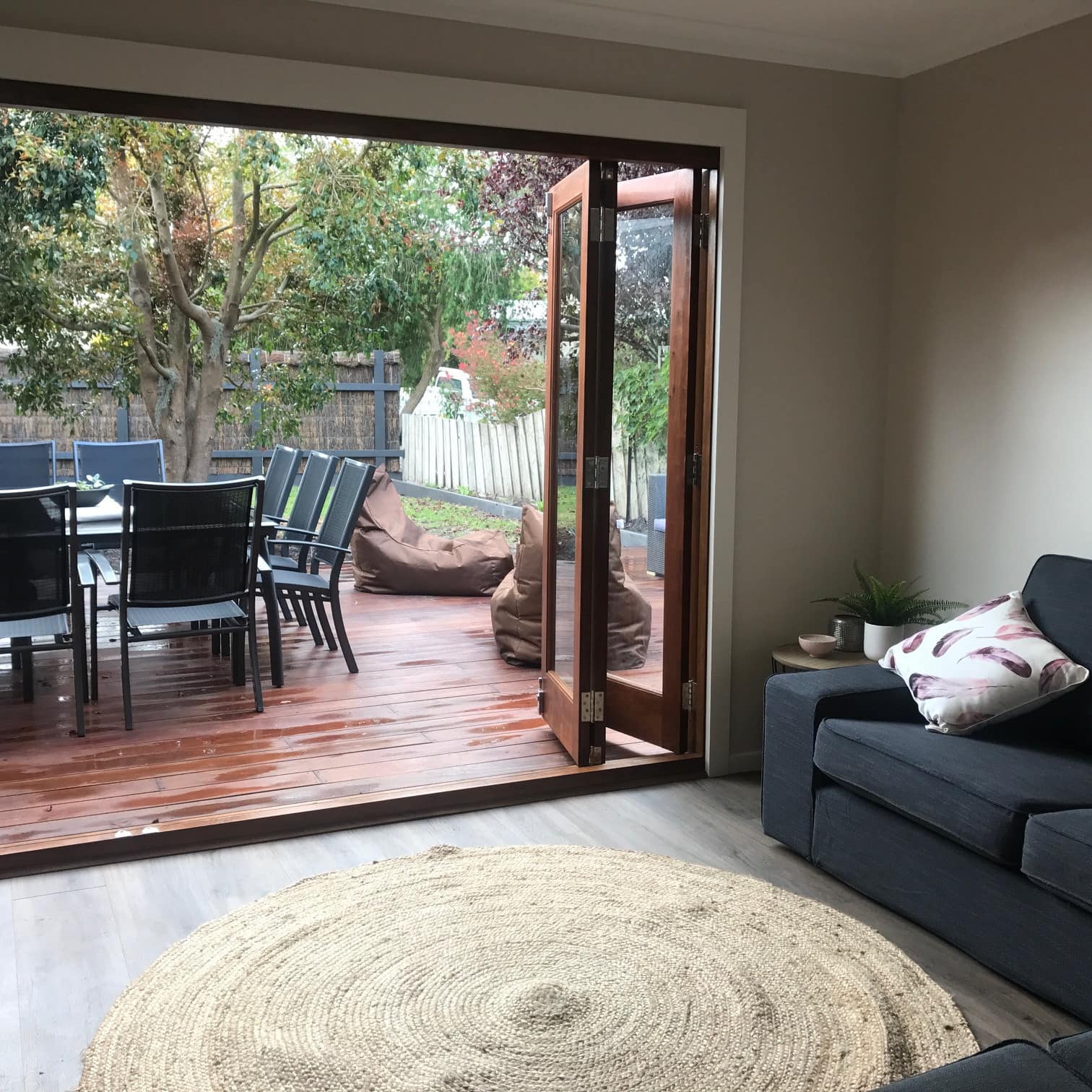Dining patio table and chairs on backyard deck viewed from opened bifold doors of a living room with a couch and area rug