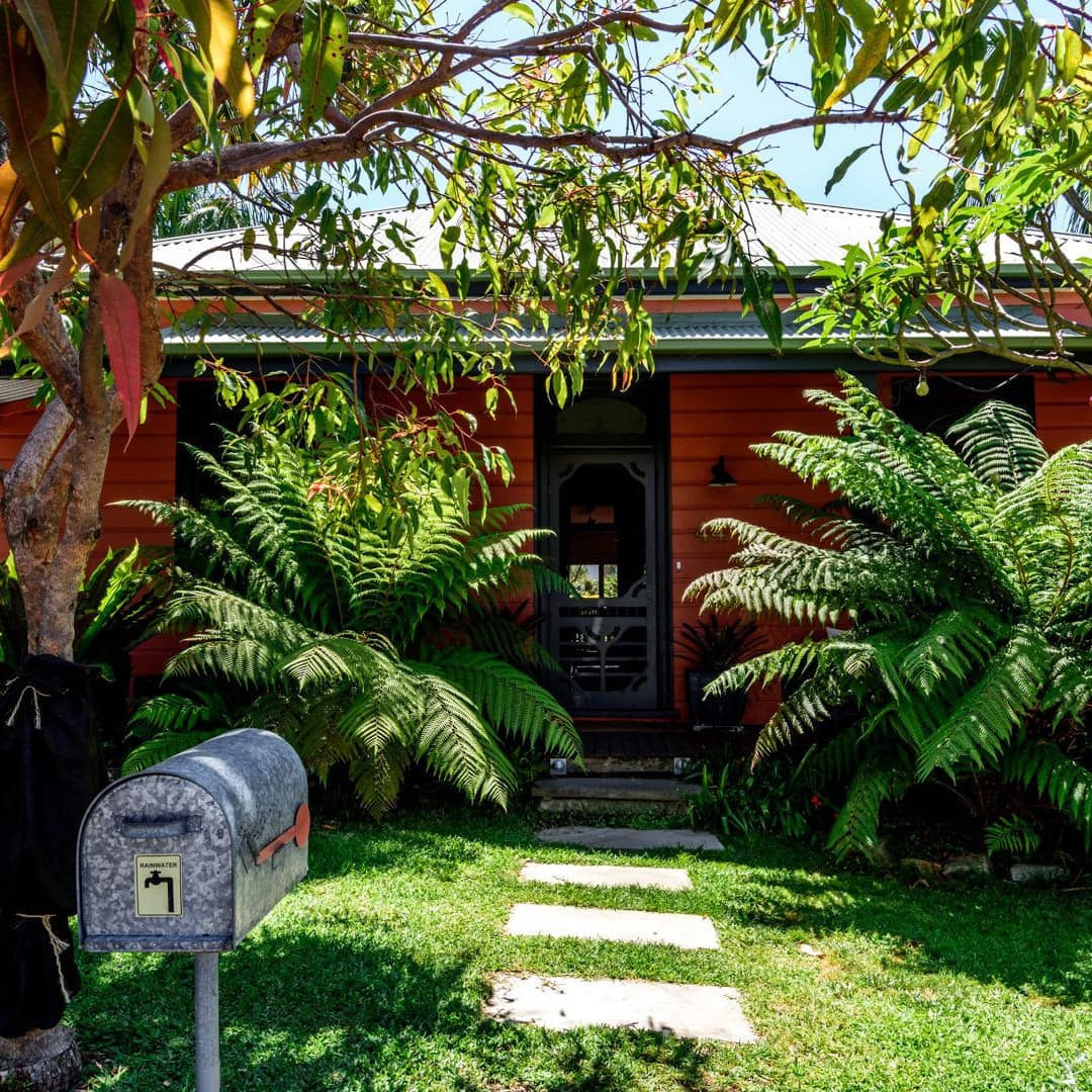 Charming red cottage surrounded by a lush front lawn with tropical plants.