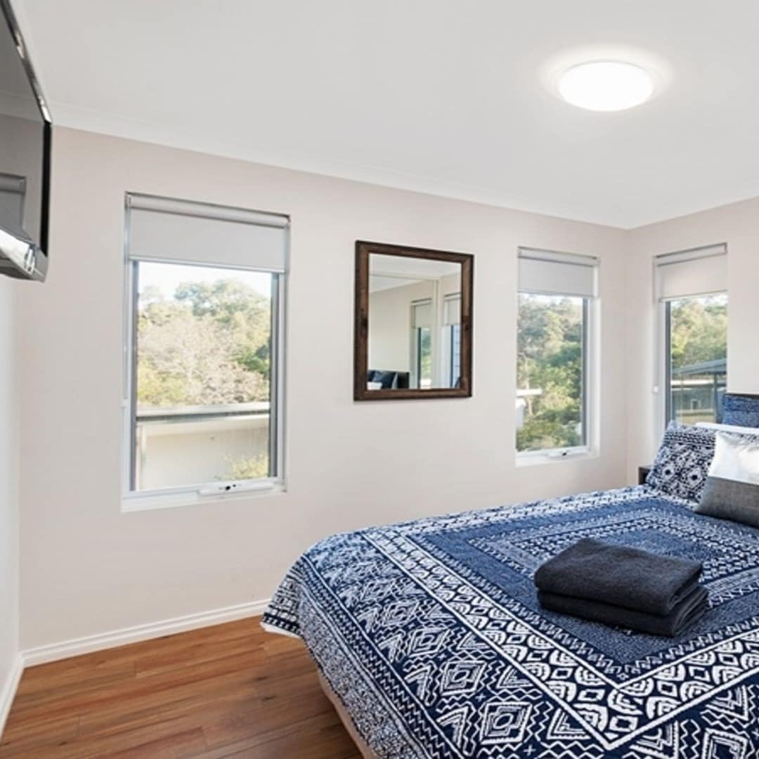 Bright bedroom with hardwood floors, white walls, a king size bed with blue and white bedding, a flatscreen TV.