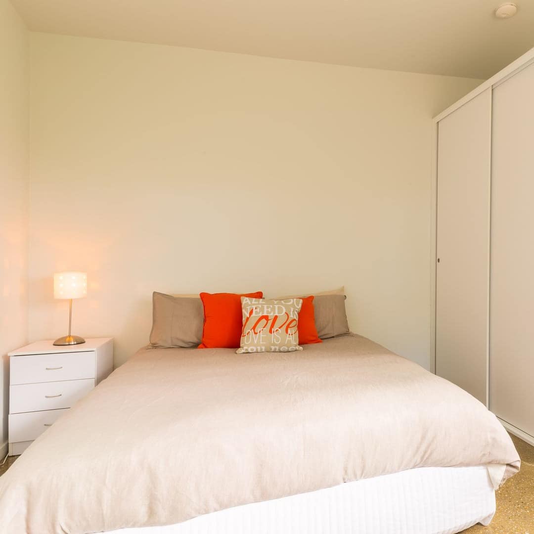 Spacious bedroom with hardwood floors, a king-size bed with tan and red bedding, a white bedside table and wardrobe.