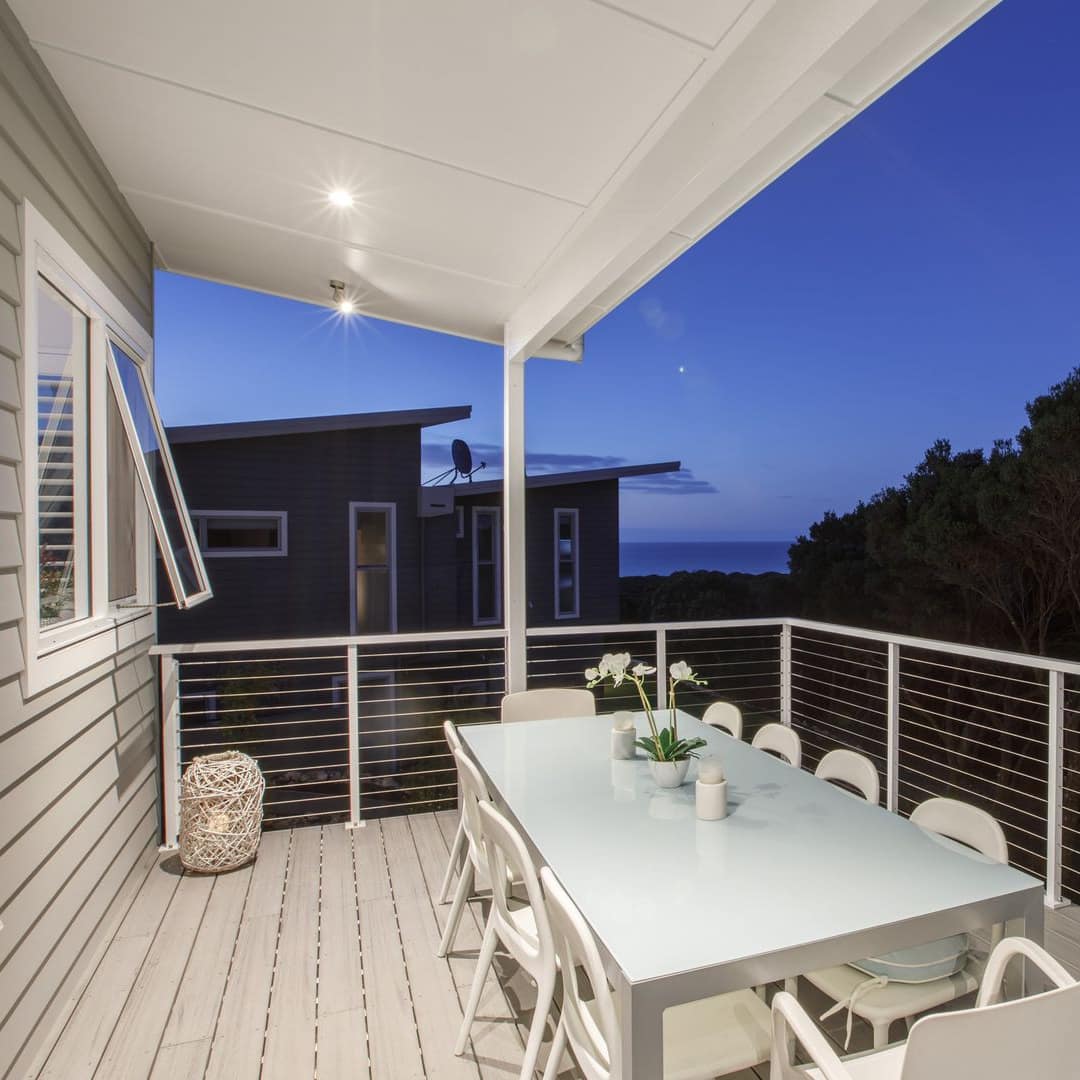 A nighttime view of a covered balcony with a white dining set overlooking a thick forested area.