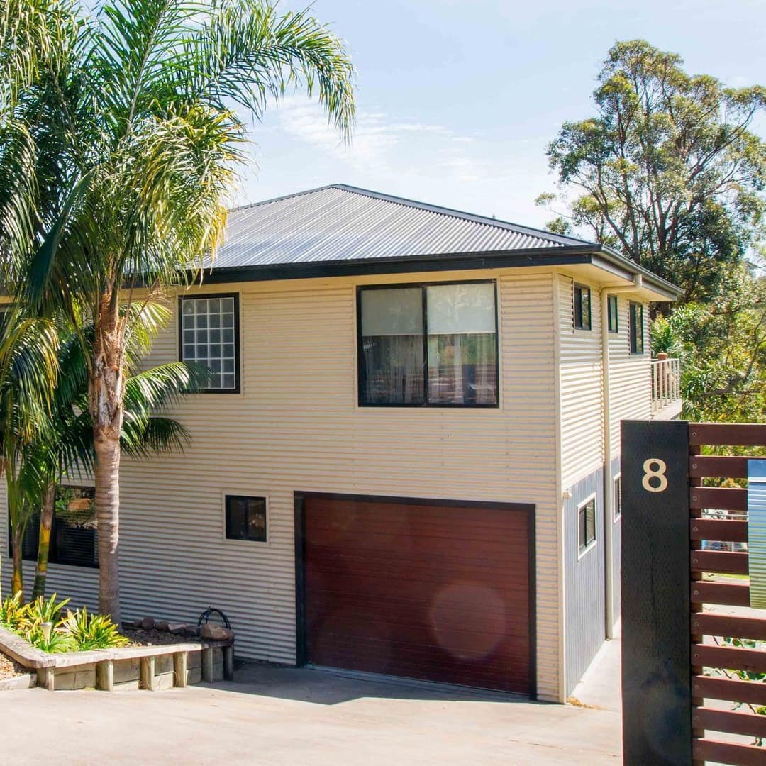 Two story home with beige vinyl siding, black roof, garage, wood slat fence, and landscaped front lawn with palm trees.