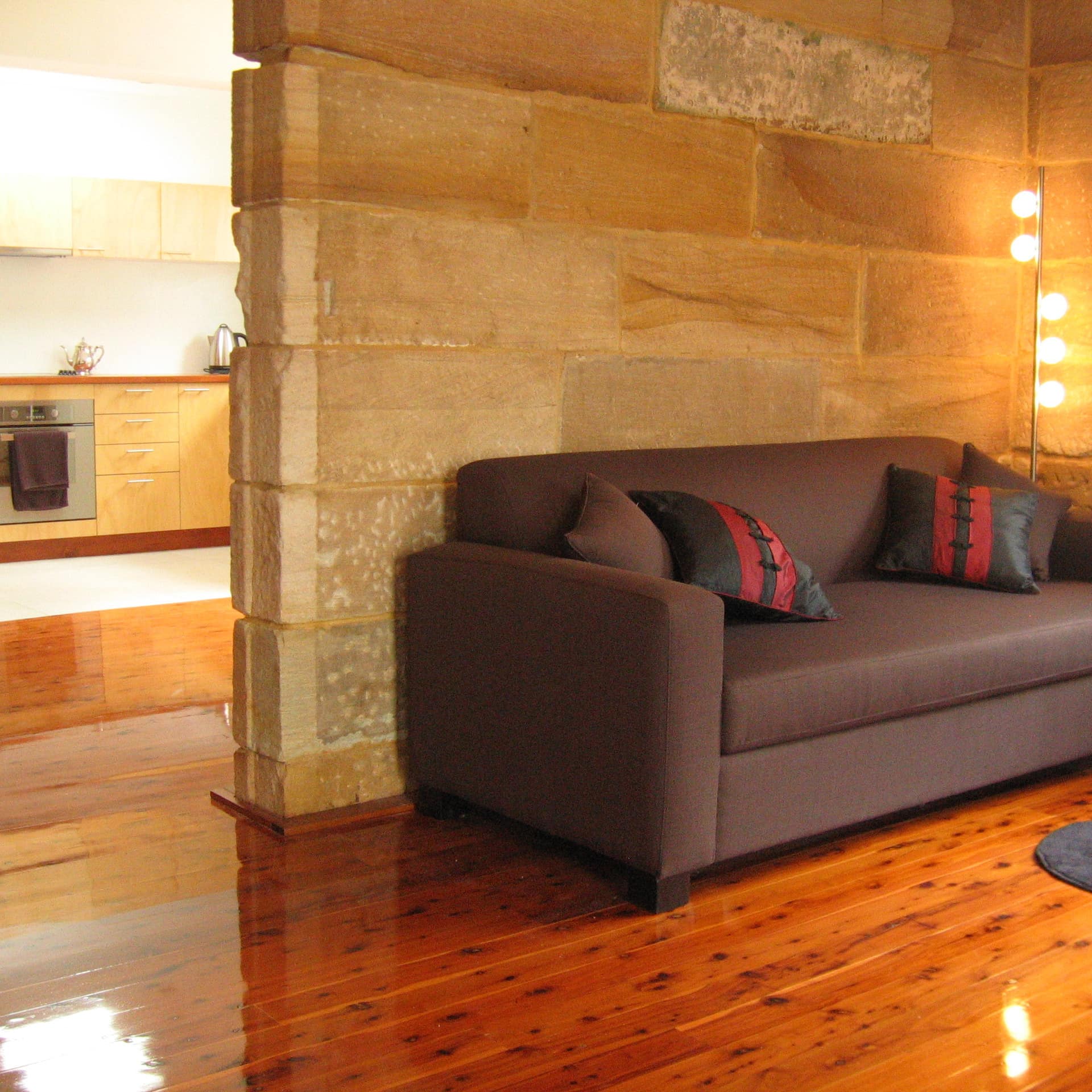 The warm interior of a cottage with high-gloss hardwood floors, sandstone walls, a brown sofa and a view of the kitchen.
