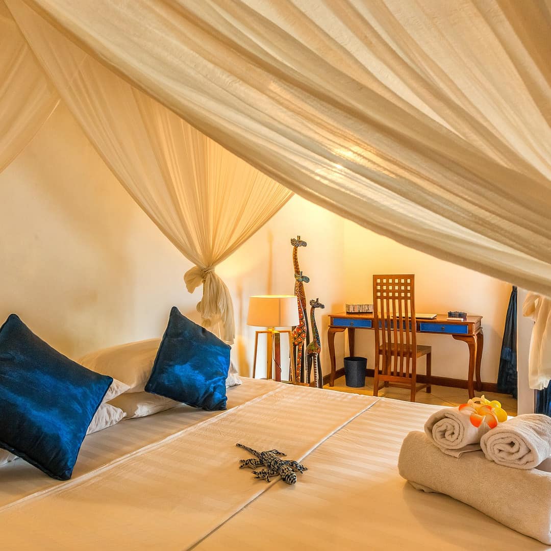 Master bedroom of a villa with a curtained bed and cream-coloured linens, royal blue accent pillows and a small writing desk