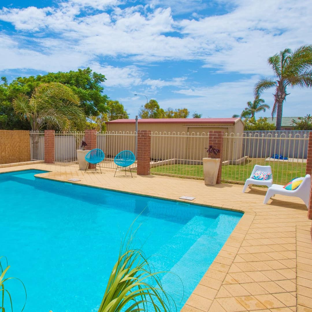 Sunny view of a pool with beige tile pool deck, plant beds, and white and blue lounge chairs enclosed by brick fence.