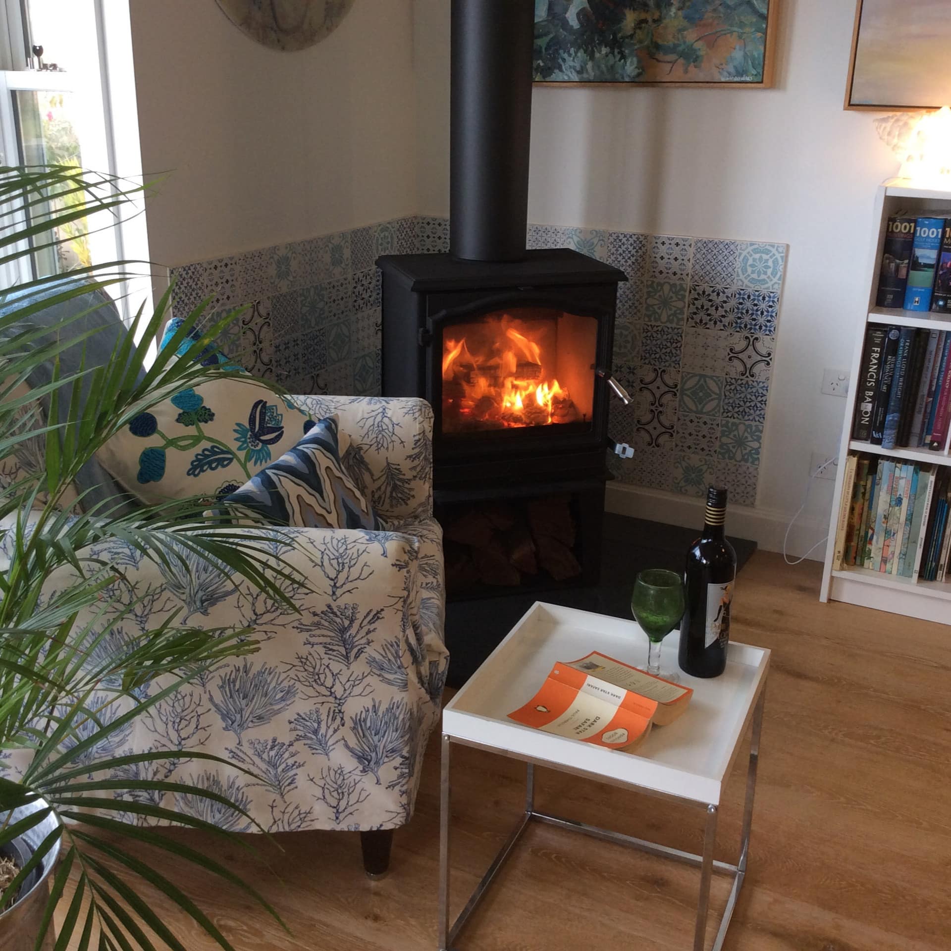 Living room with blue and white print chair, wood-burning stove, coffee table with wine, full bookshelf, and a potted plant.