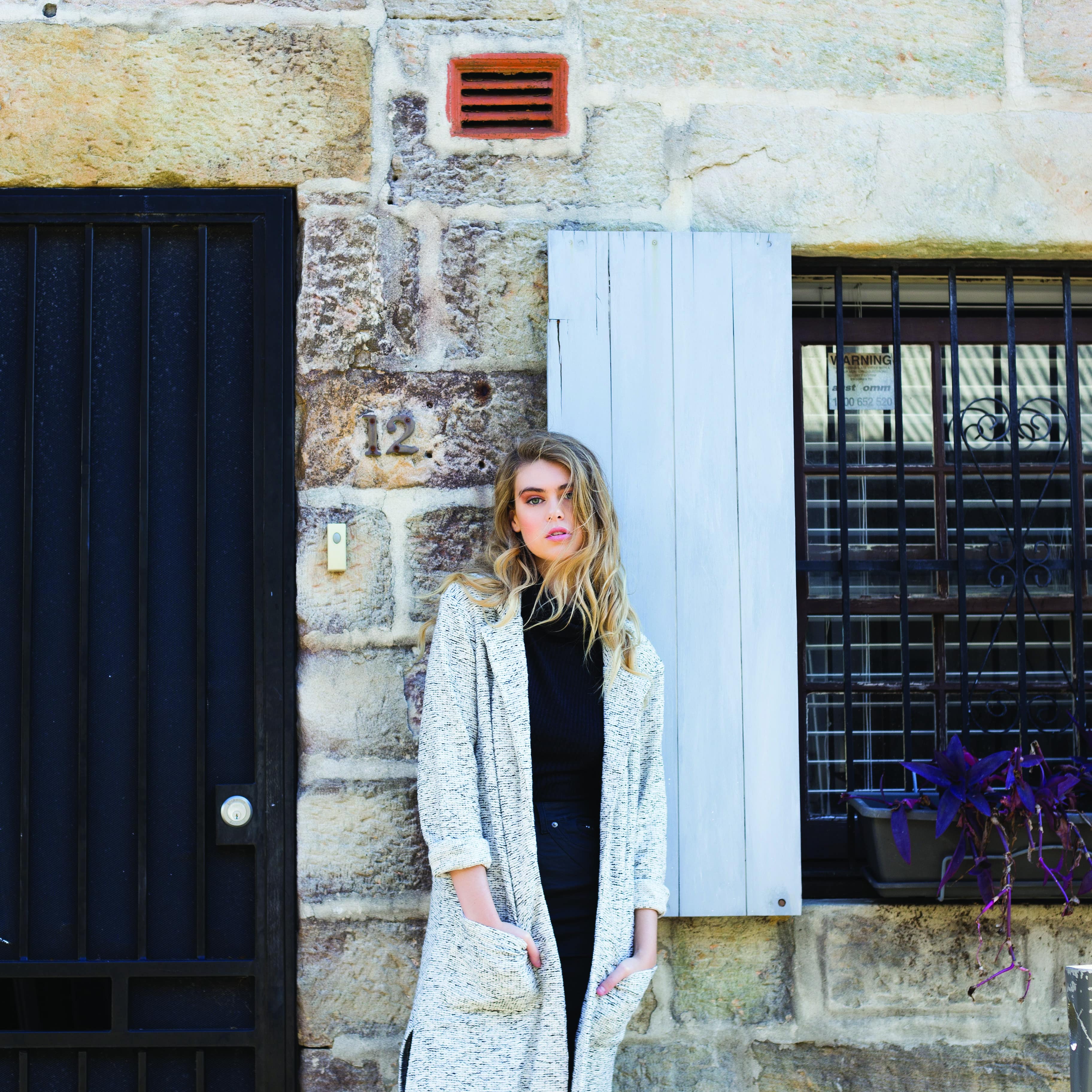 Young woman with long blonde hair in a grey sweater stands in front of sandstone cottage with black door